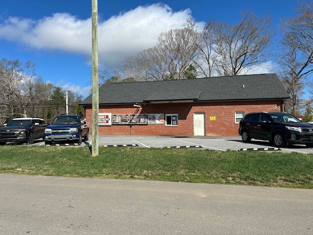 A single-story red brick building with a dark shingled roof, a parking lot, and three parked cars on a sunny day.