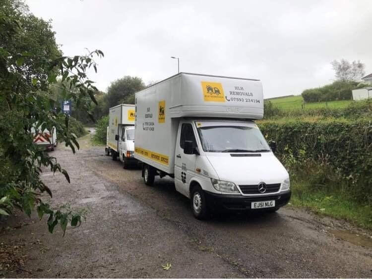 Two white moving vans parked on a narrow road; overcast sky; foliage on either side.