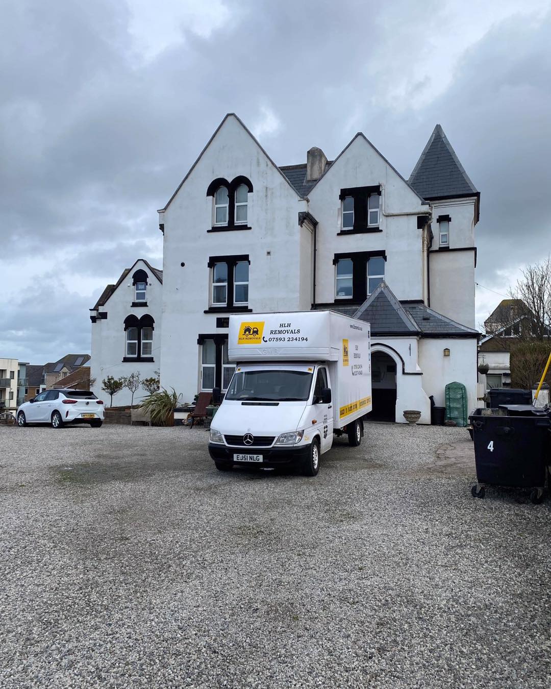 White moving van parked in front of a large white building with multiple windows. Cloudy day.