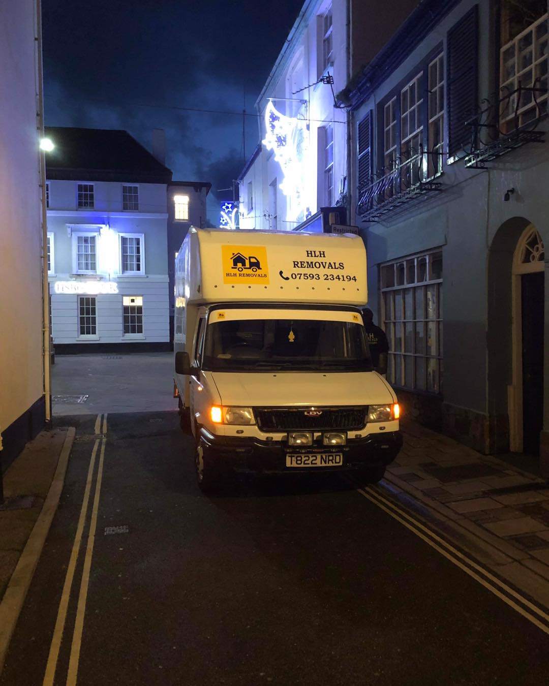 A removals van parked on a narrow street at night, lit by streetlights, with buildings on either side.