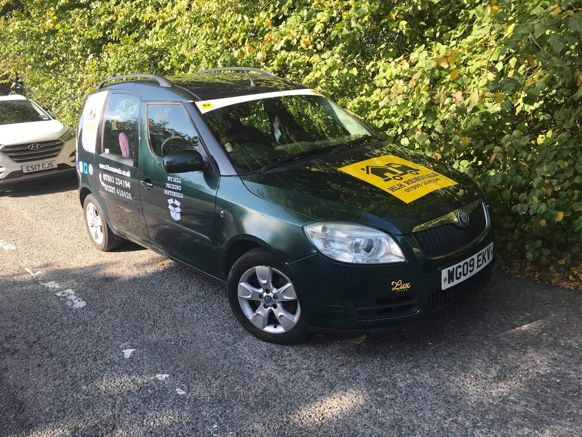 Green Skoda car parked next to bushes, with business logo on hood, and a white car partially visible.