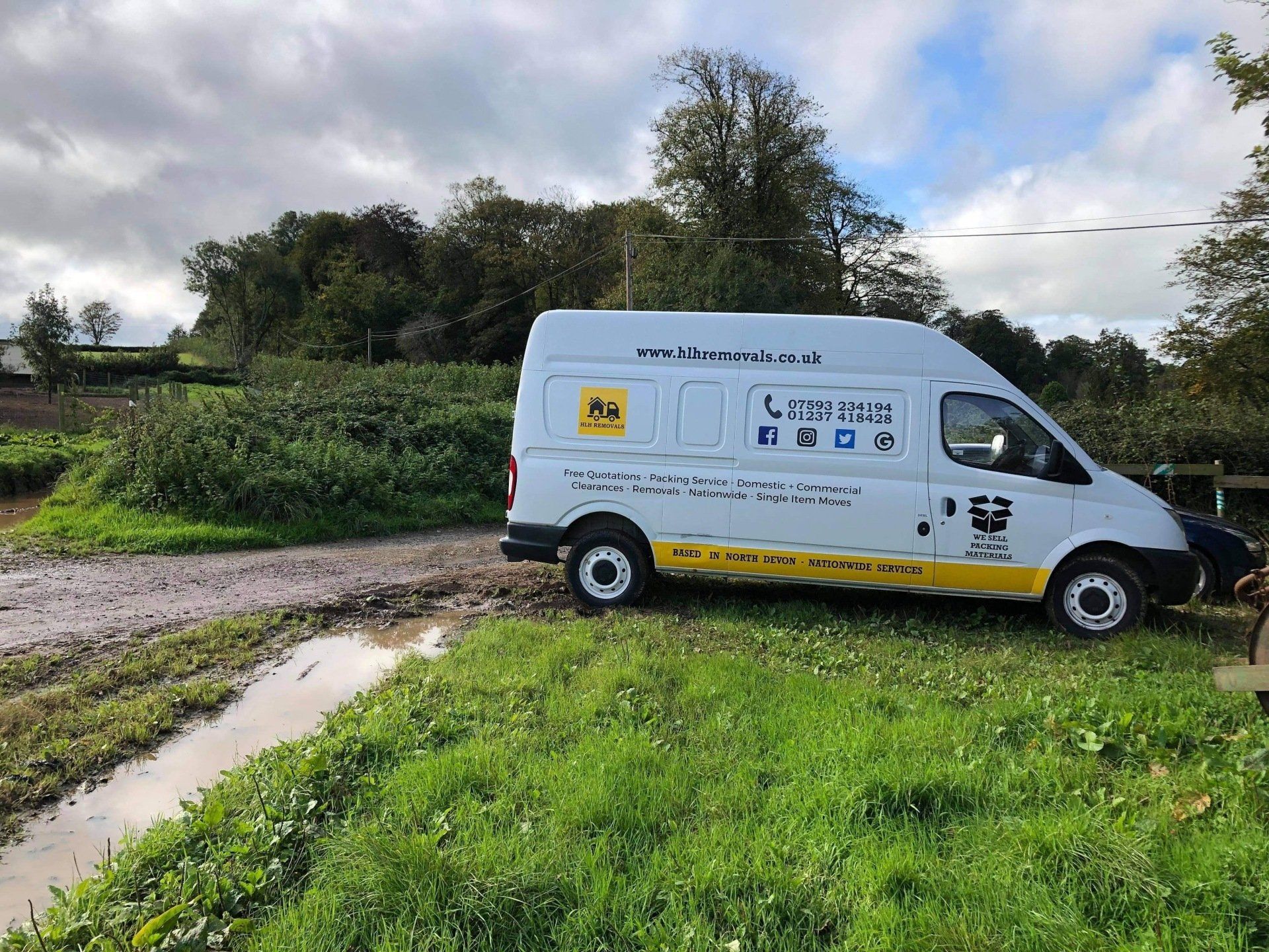 White service van parked on muddy rural road, near a field of plants. Overcast sky.