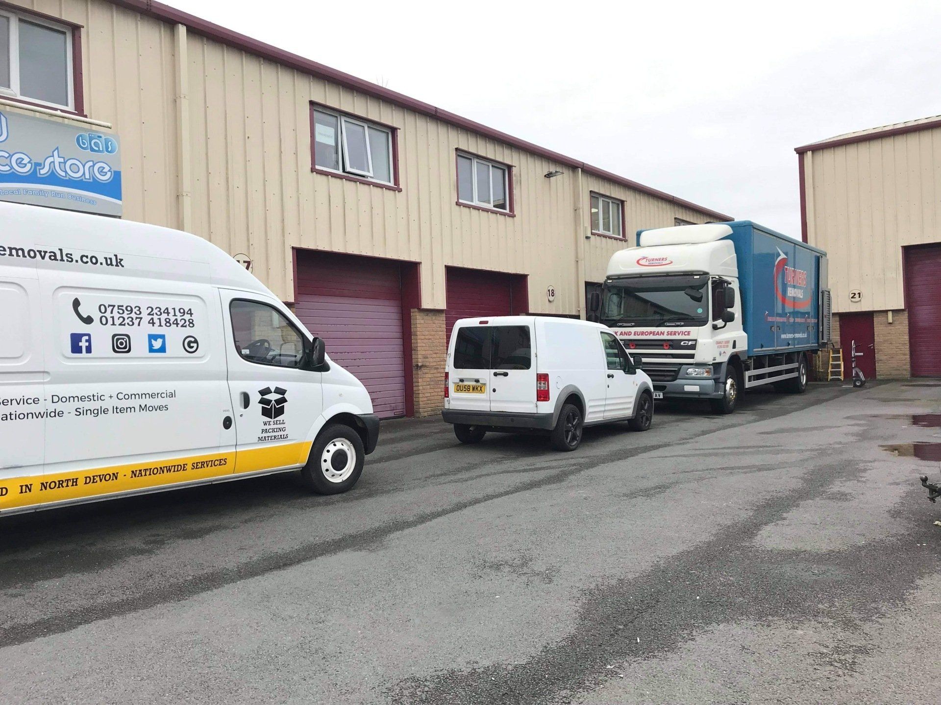 Three vehicles parked outside a commercial building: a white van, a white car, and a blue and white semi-truck.