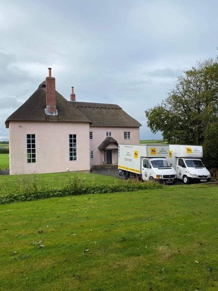 Pink cottage with thatched roof and two moving trucks parked on the lawn.
