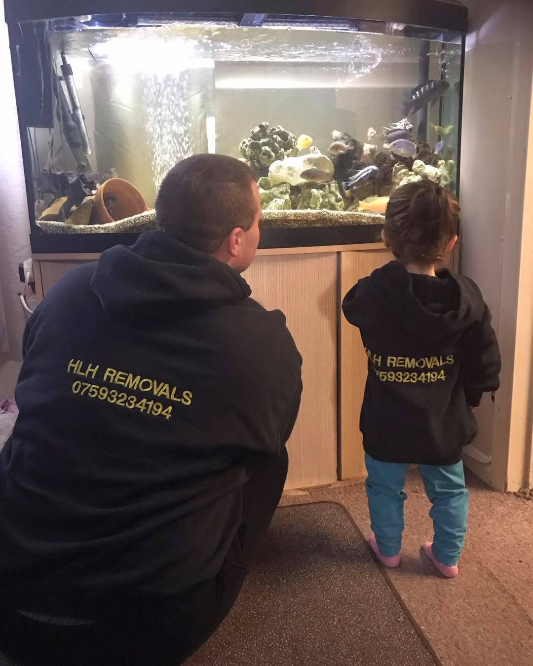 Father and child wearing matching black hoodies, looking at fish tank. Light brown cabinet beneath.