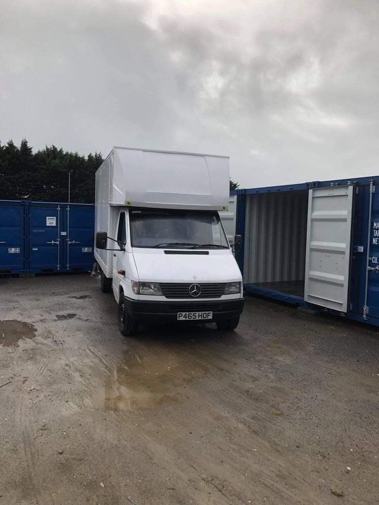 White box truck parked between blue storage containers on a wet, grey day.