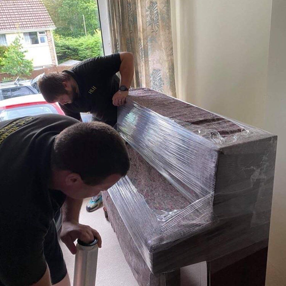 Two men wrap a piano in plastic wrap near a window.