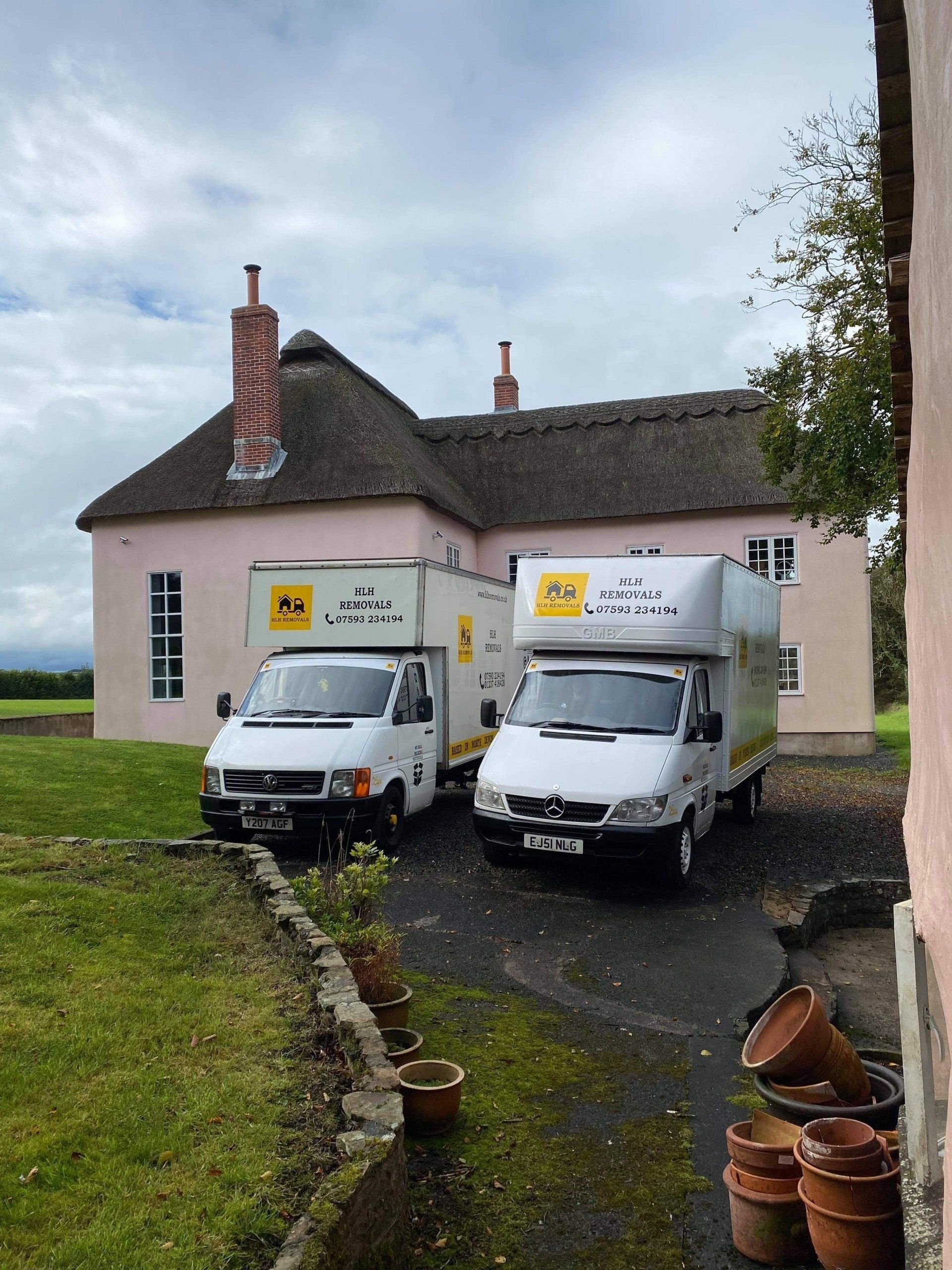 Two white moving vans parked in front of a pink thatched-roof house.