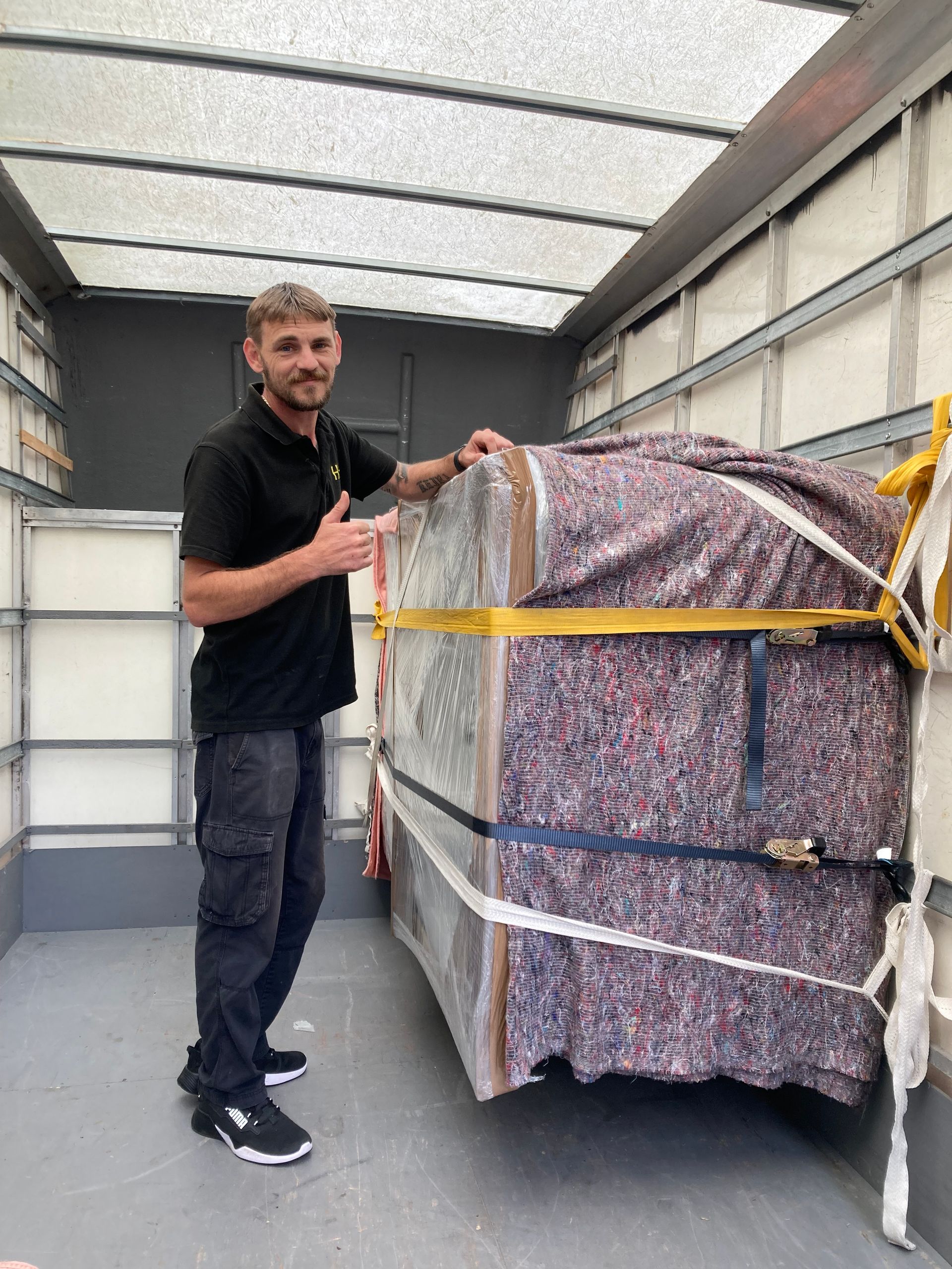 Man in black shirt gives thumbs up, standing next to a large, wrapped item inside a truck.