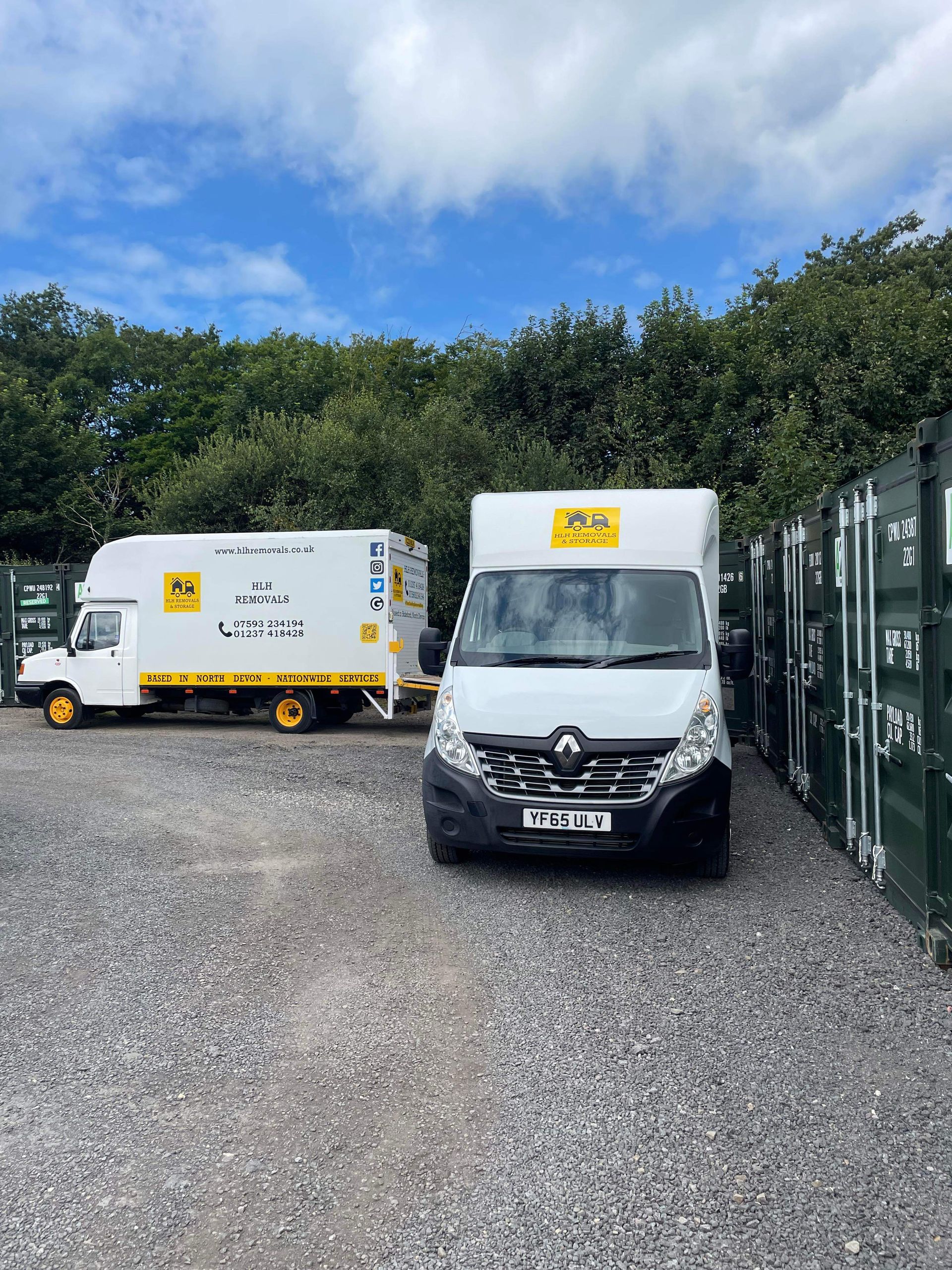 Two white vans with yellow accents parked on gravel, surrounded by green storage containers and trees.
