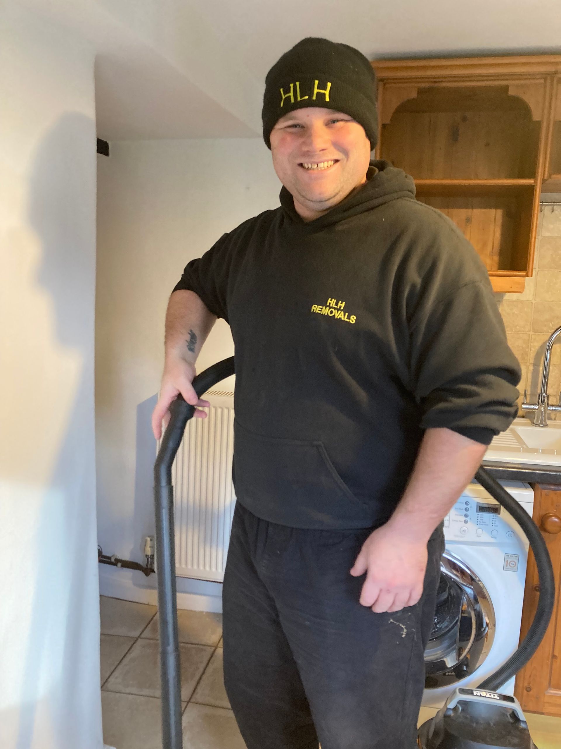 Man in black hat and sweatshirt smiles, holding a vacuum, in a kitchen.