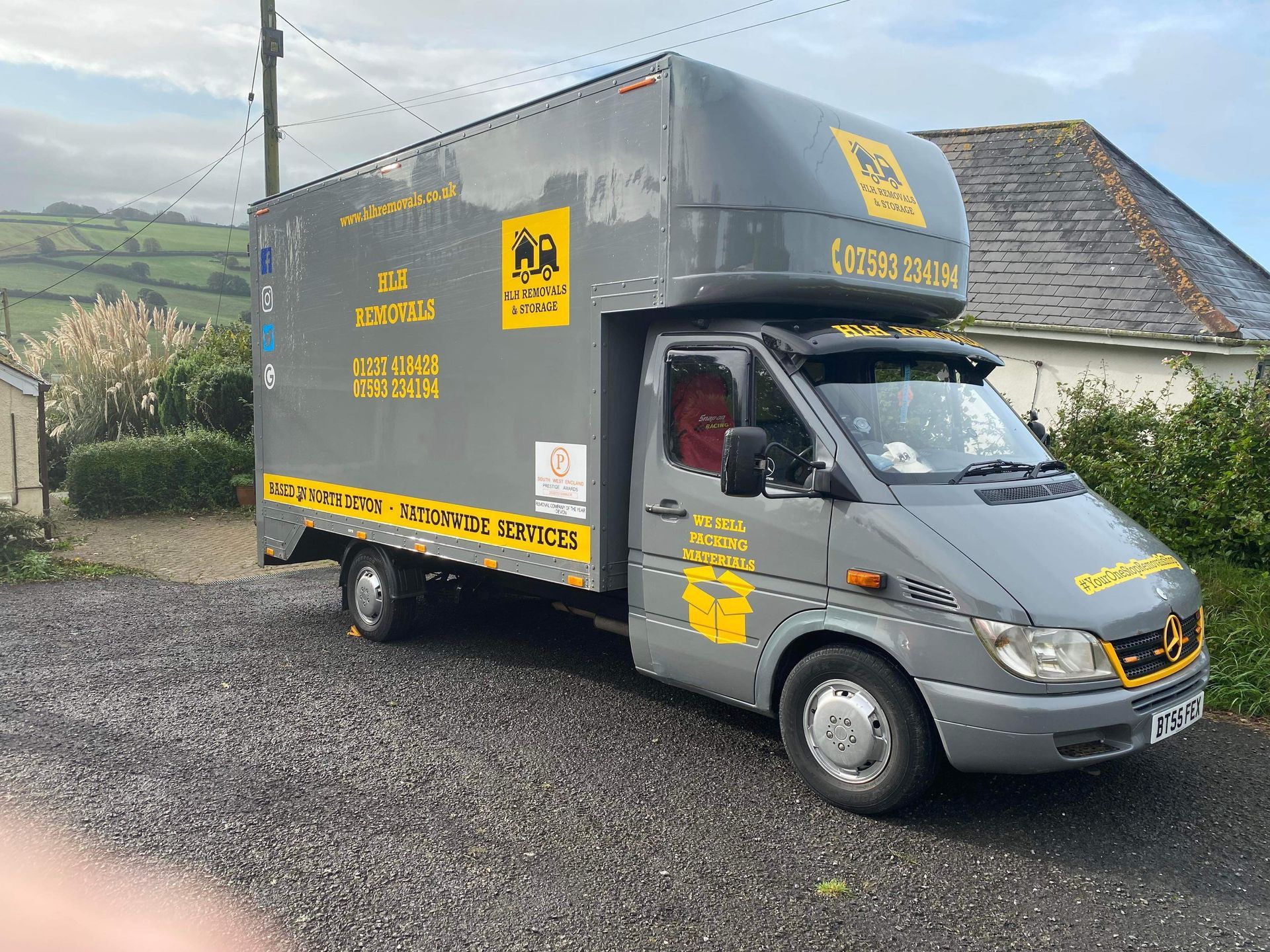 Gray delivery truck parked on gravel driveway in front of a house, overcast sky.