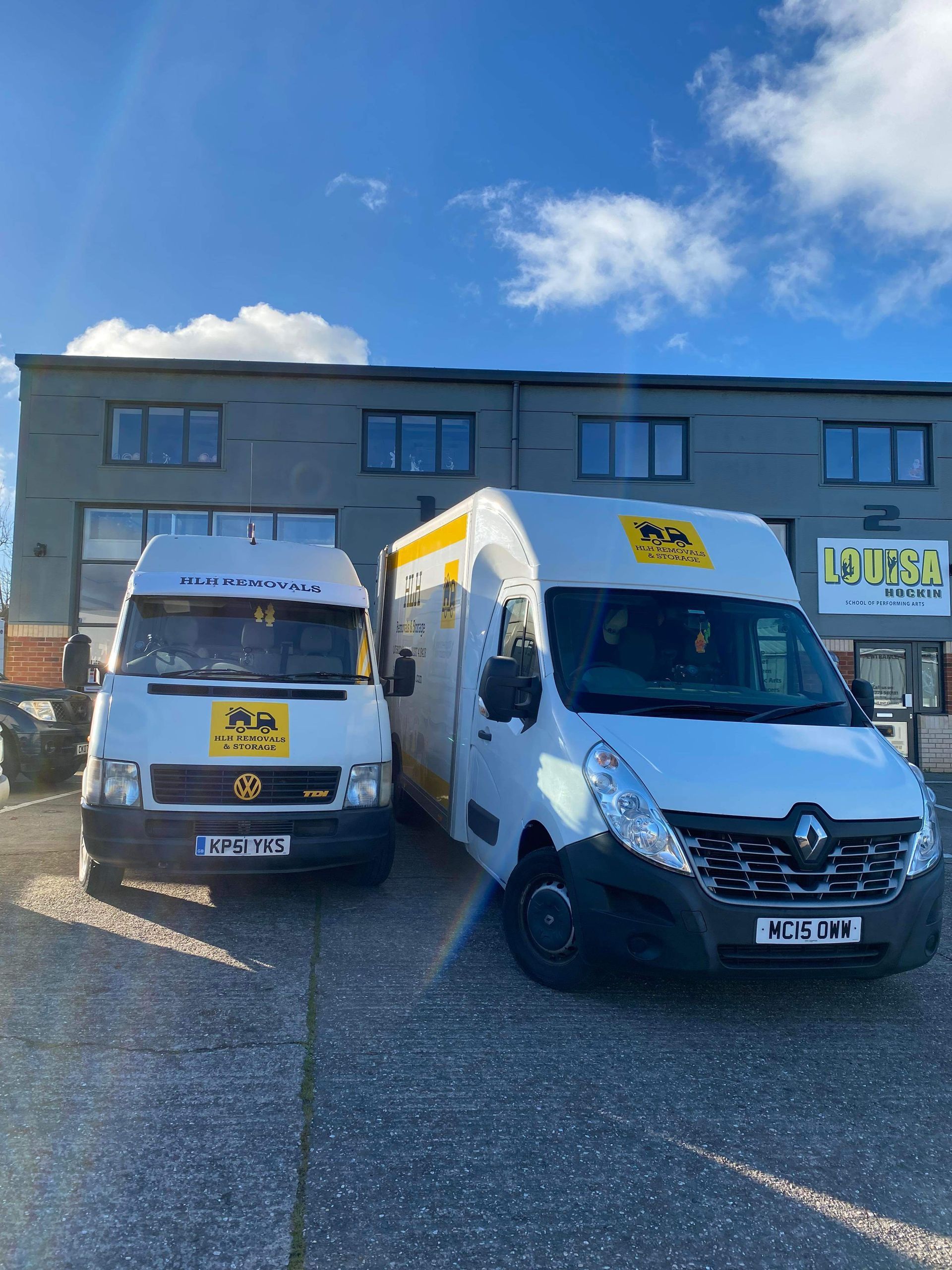 Two white delivery vans parked in front of a gray building on a sunny day.