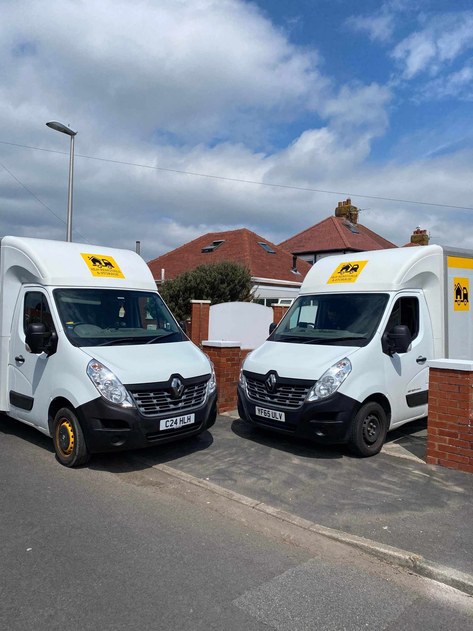 Two white moving vans with yellow logos parked on a residential street, under a partly cloudy sky.