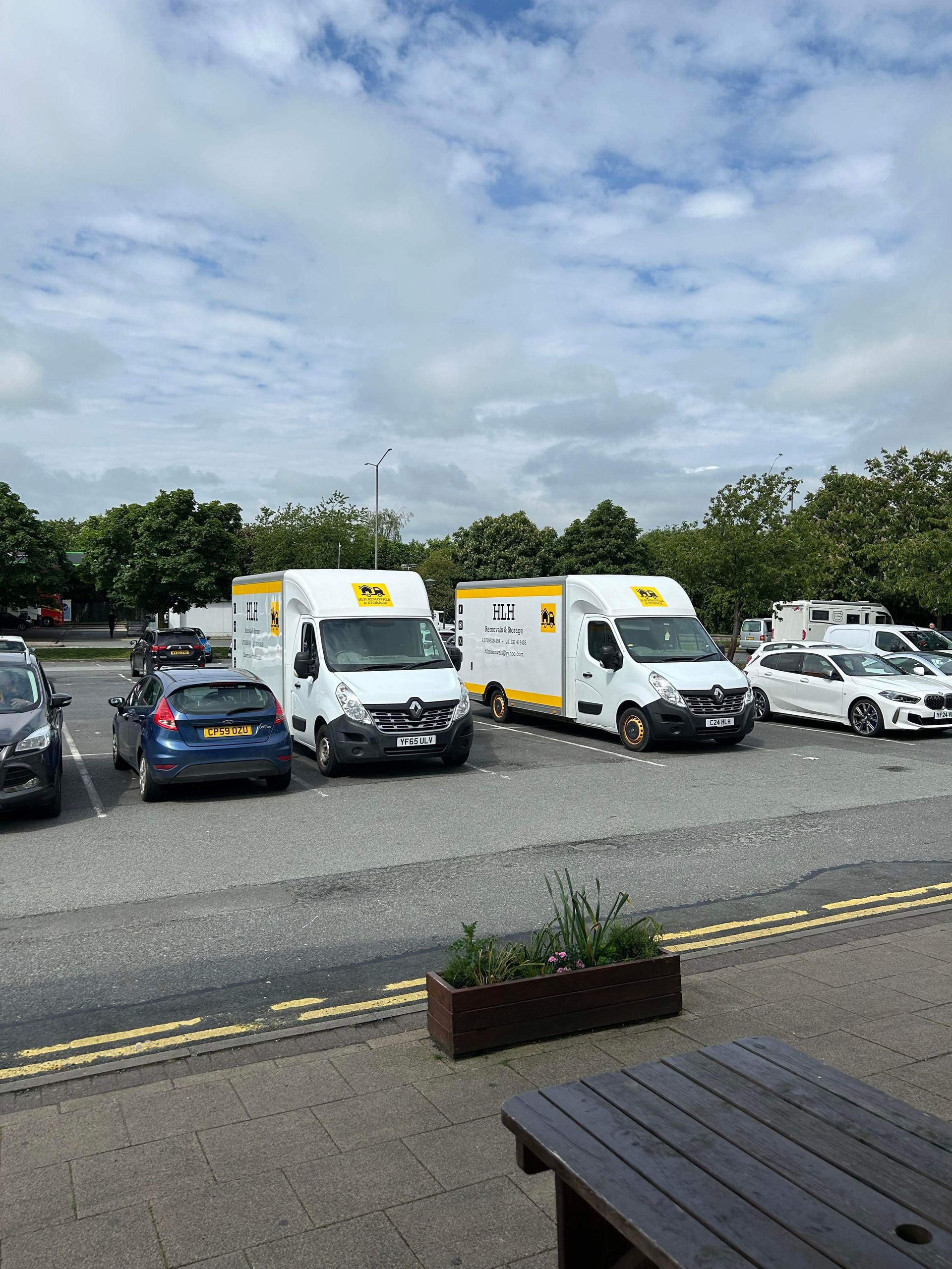 Two white vans with yellow logos parked in a lot with other vehicles on a cloudy day.