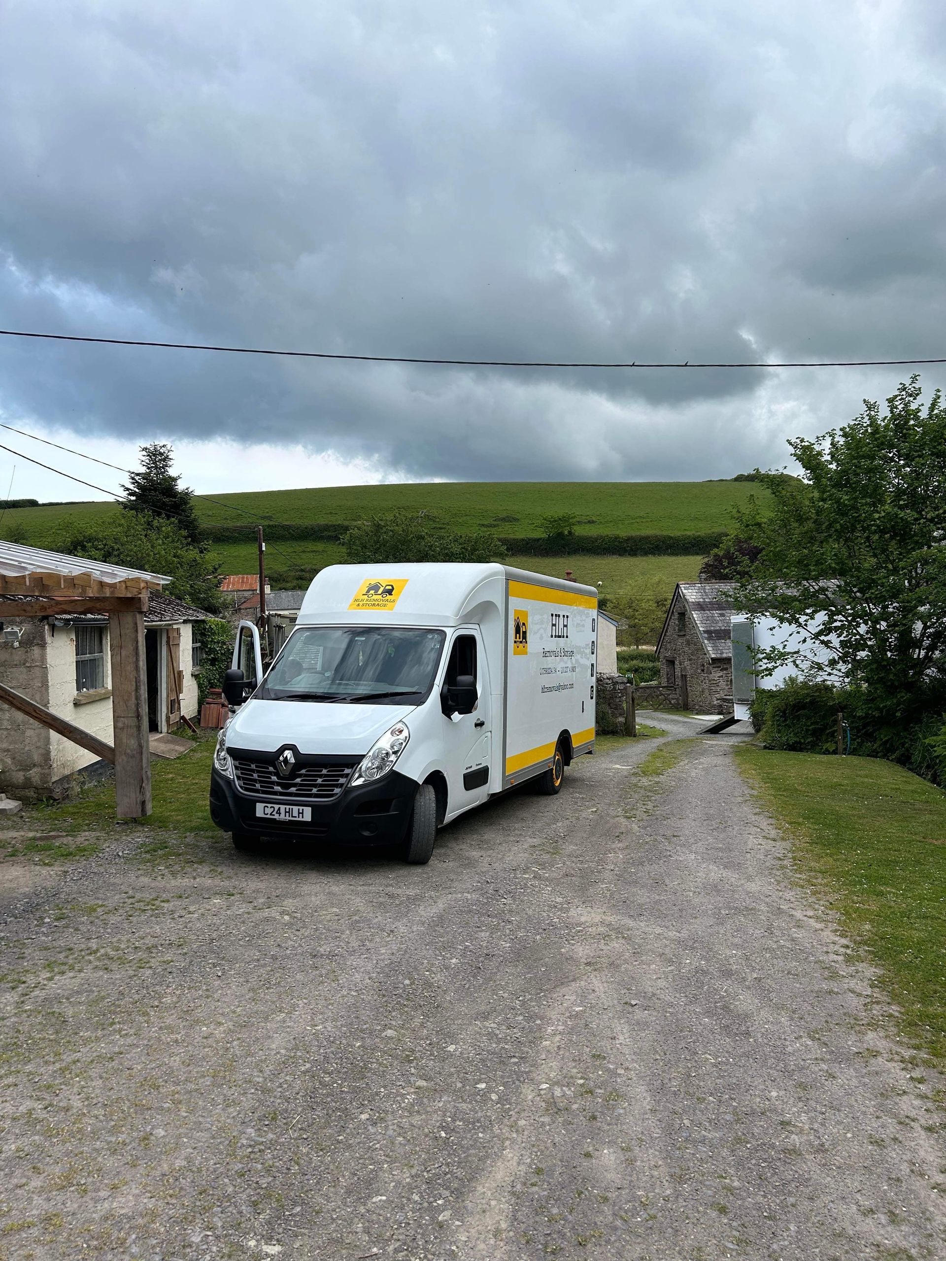 White delivery van parked on a gravel drive in front of a rural building, with a grassy hill in the background.