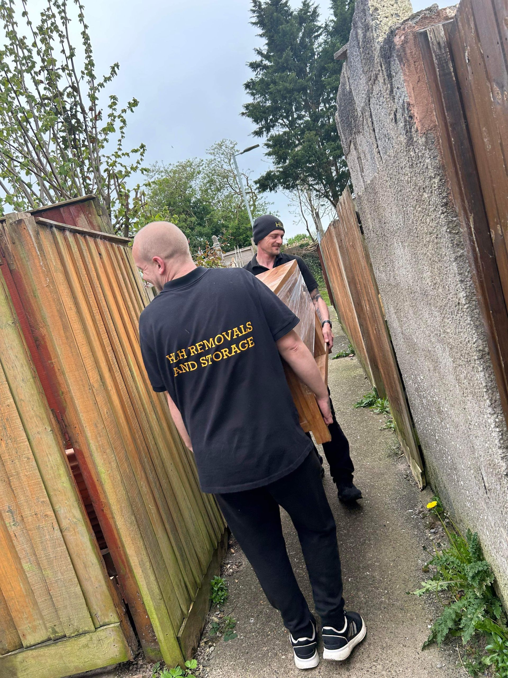 Two men carrying a large item through a narrow passageway with fences and a concrete wall.