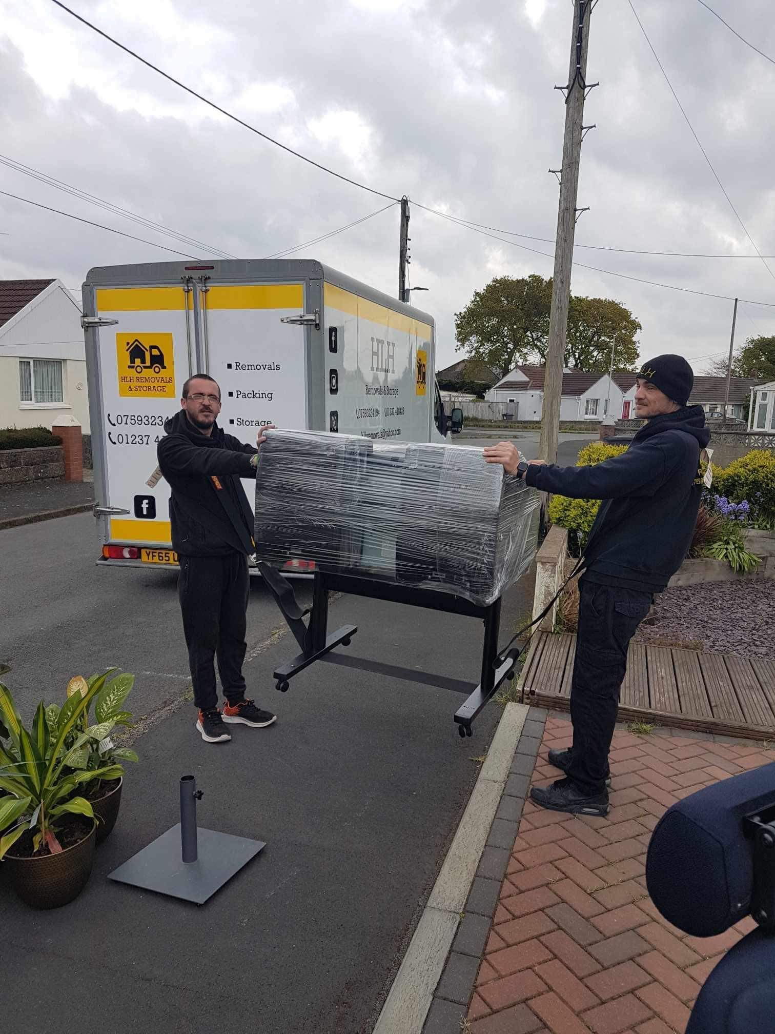 Two men carrying a large wrapped object, likely furniture, from a trailer in a residential area.