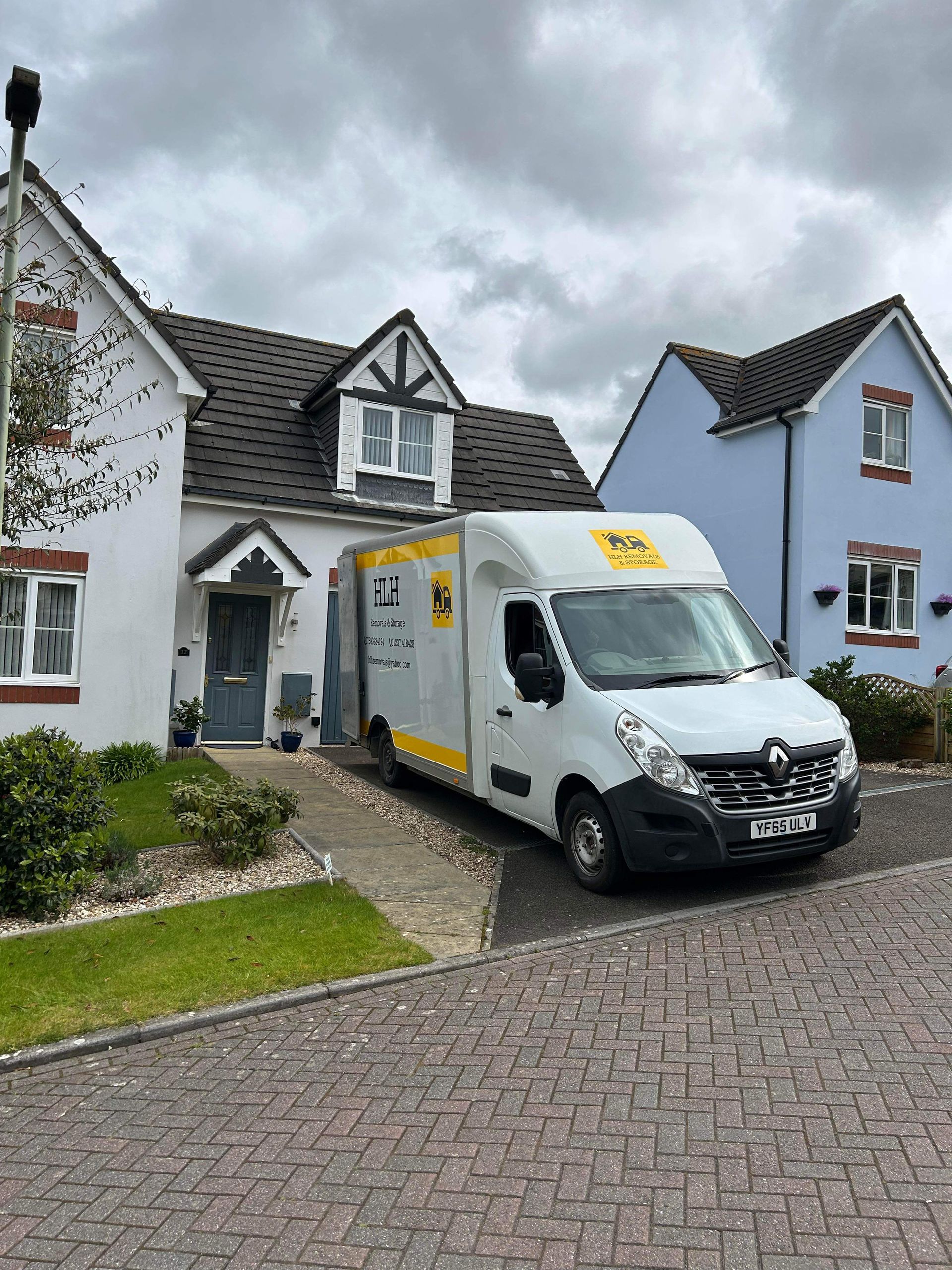 White van parked in front of a white house with a blue house to the right on a cobblestone driveway.