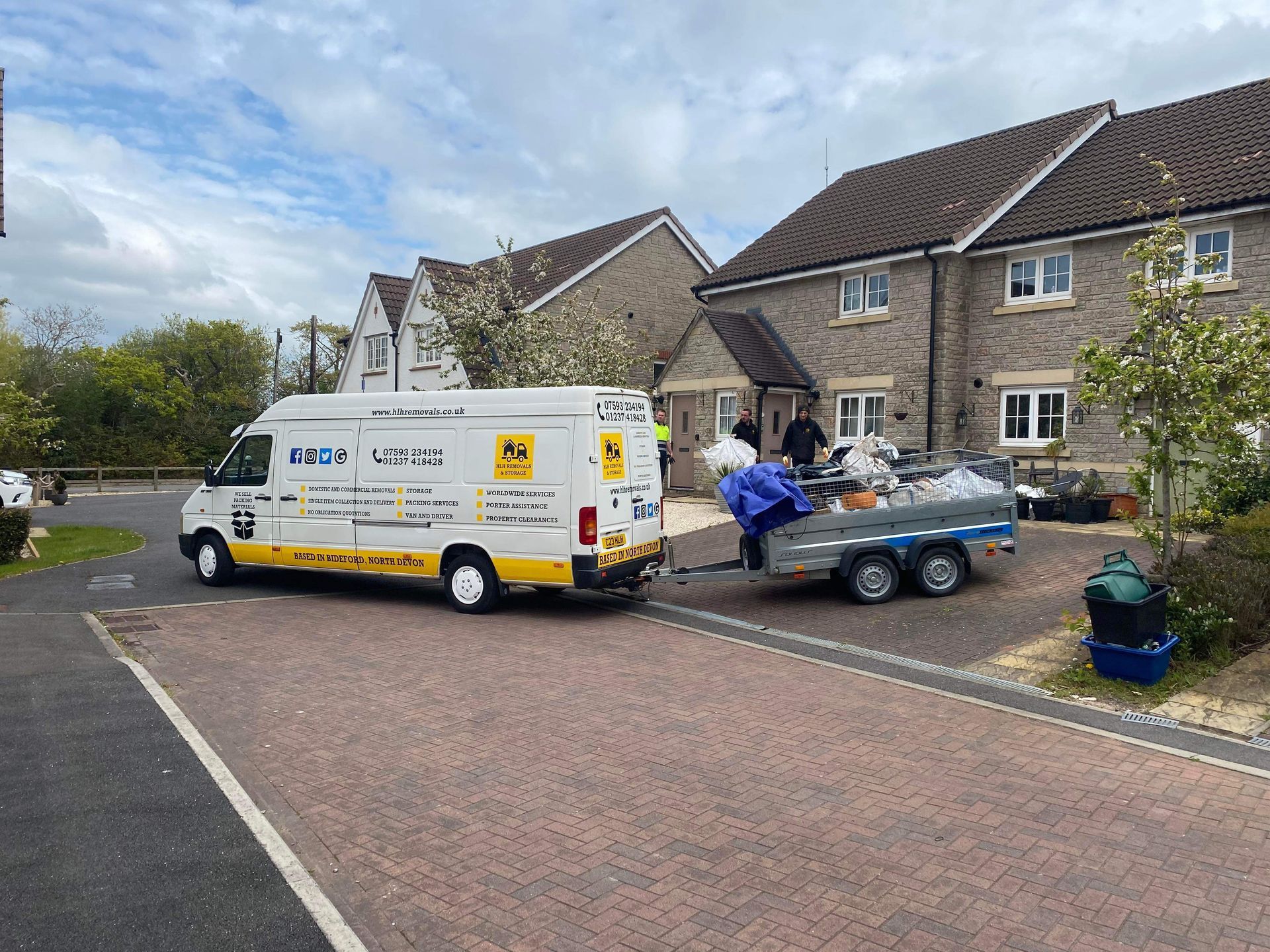 A white van with a trailer loaded with trash parked on a brick road in front of houses.