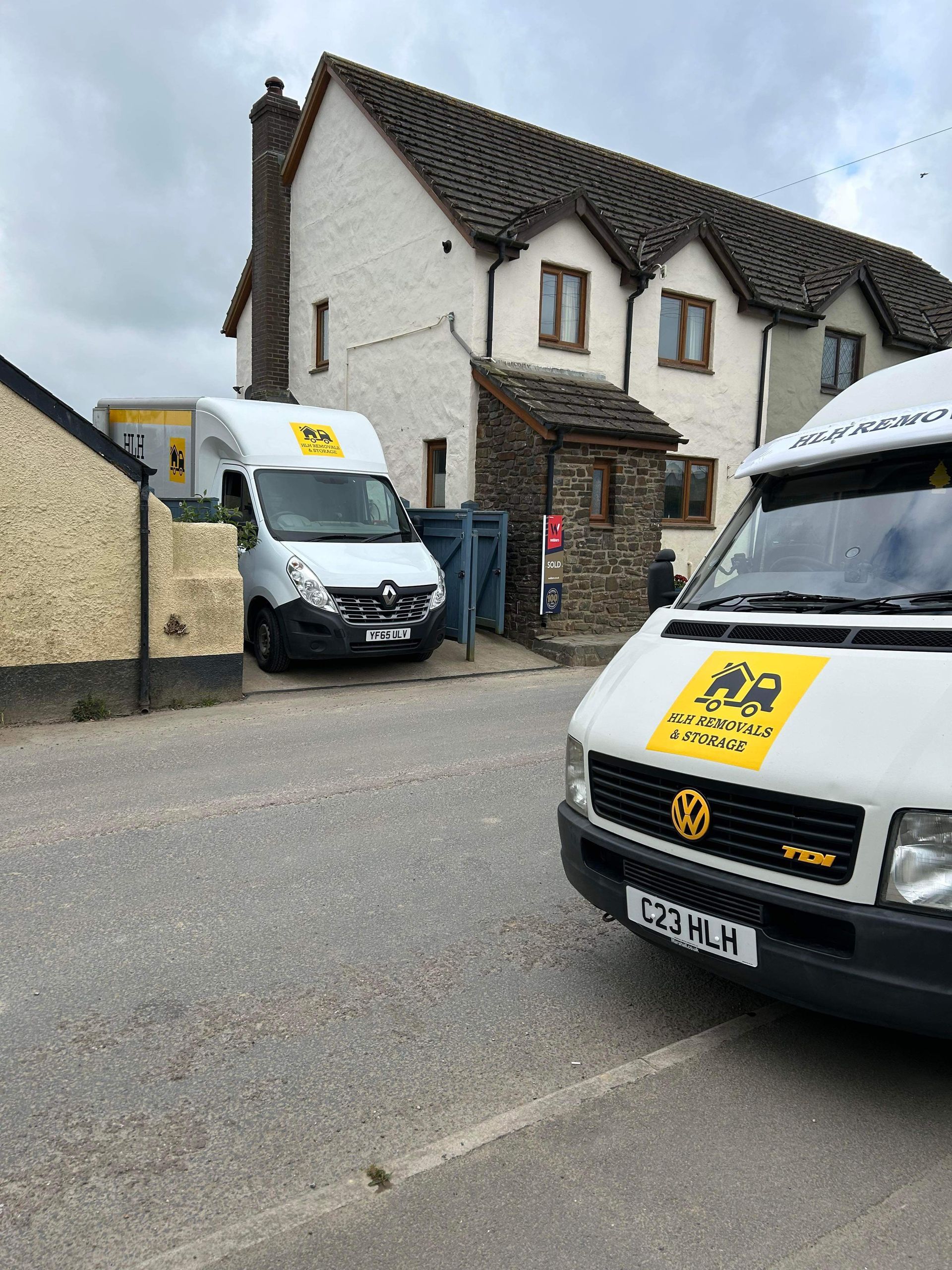 Two white vans with yellow logos parked on a street in front of a light-colored house.