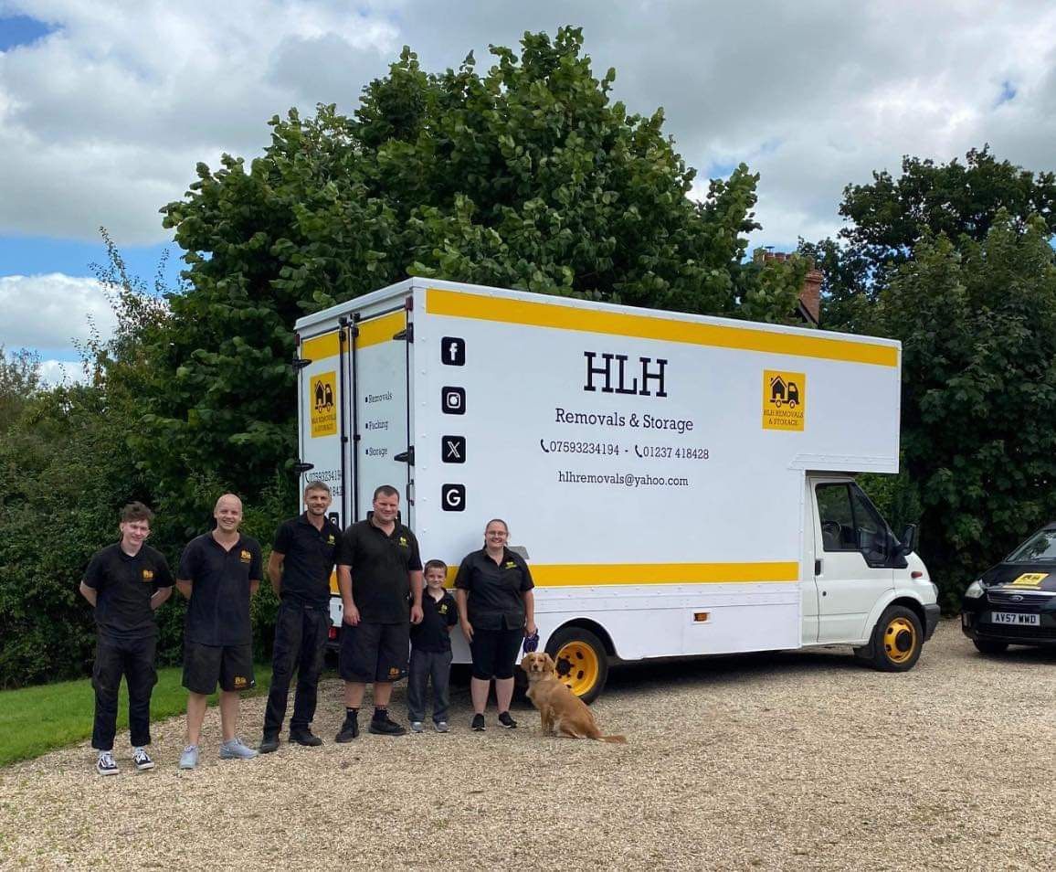 Group poses with moving van. White van with yellow accents and HLH logo, parked on gravel. People wear black shirts.