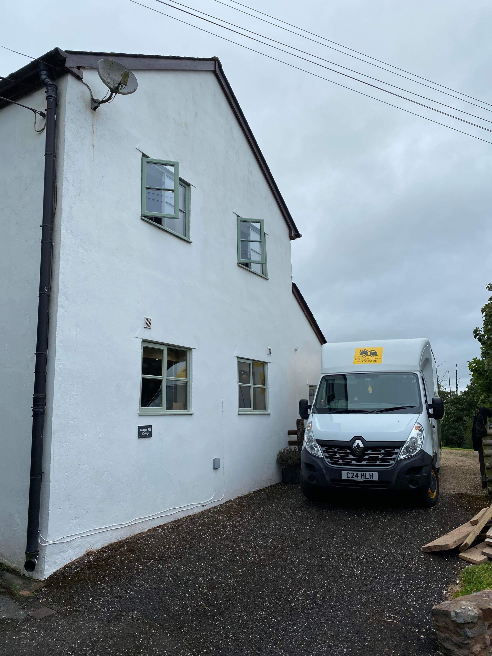 White house with green-framed windows, a white van parked in the driveway, and an overcast sky.