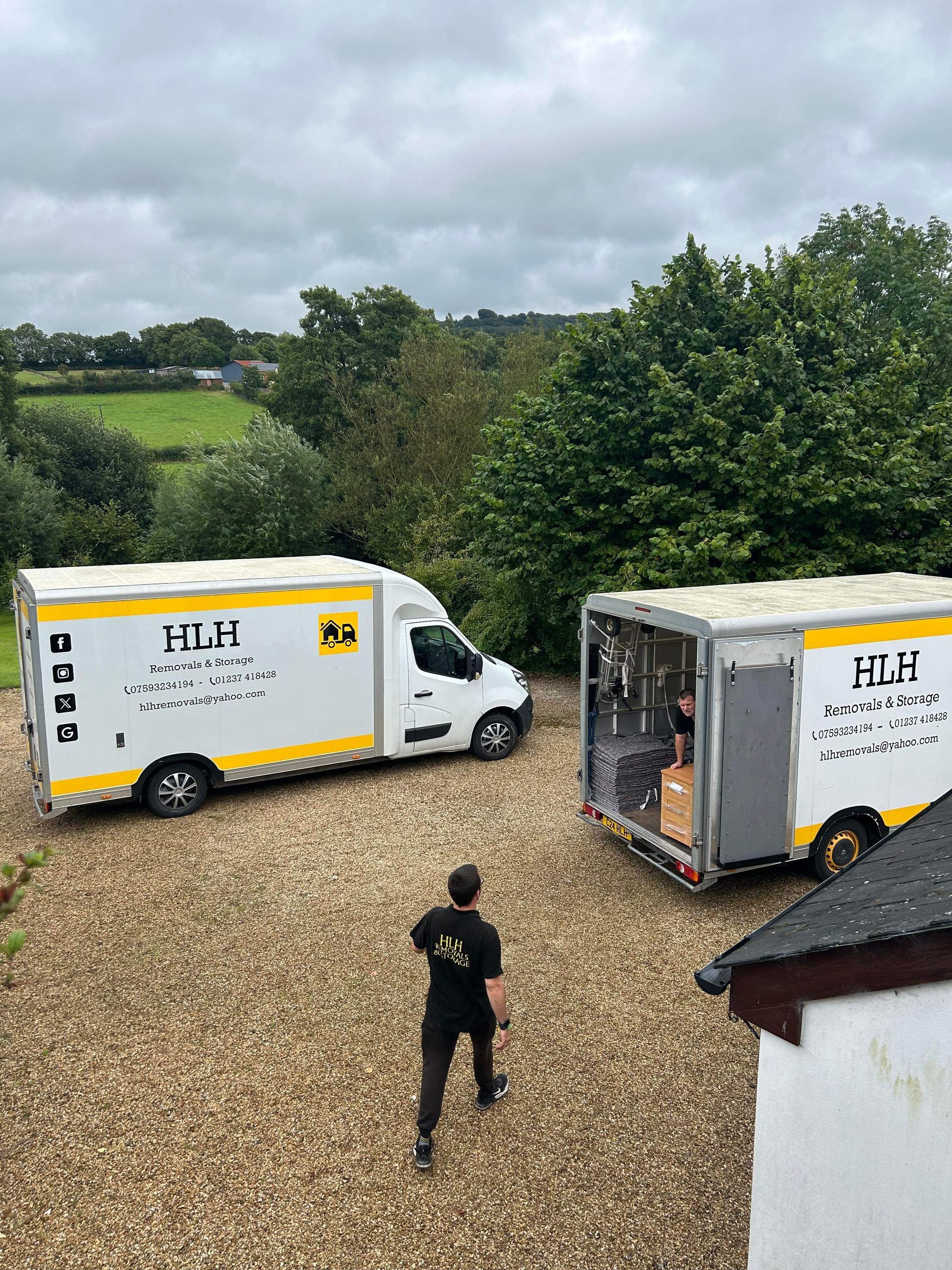 Two white HLH moving vans parked on gravel, person walking towards one, green trees and fields in background.