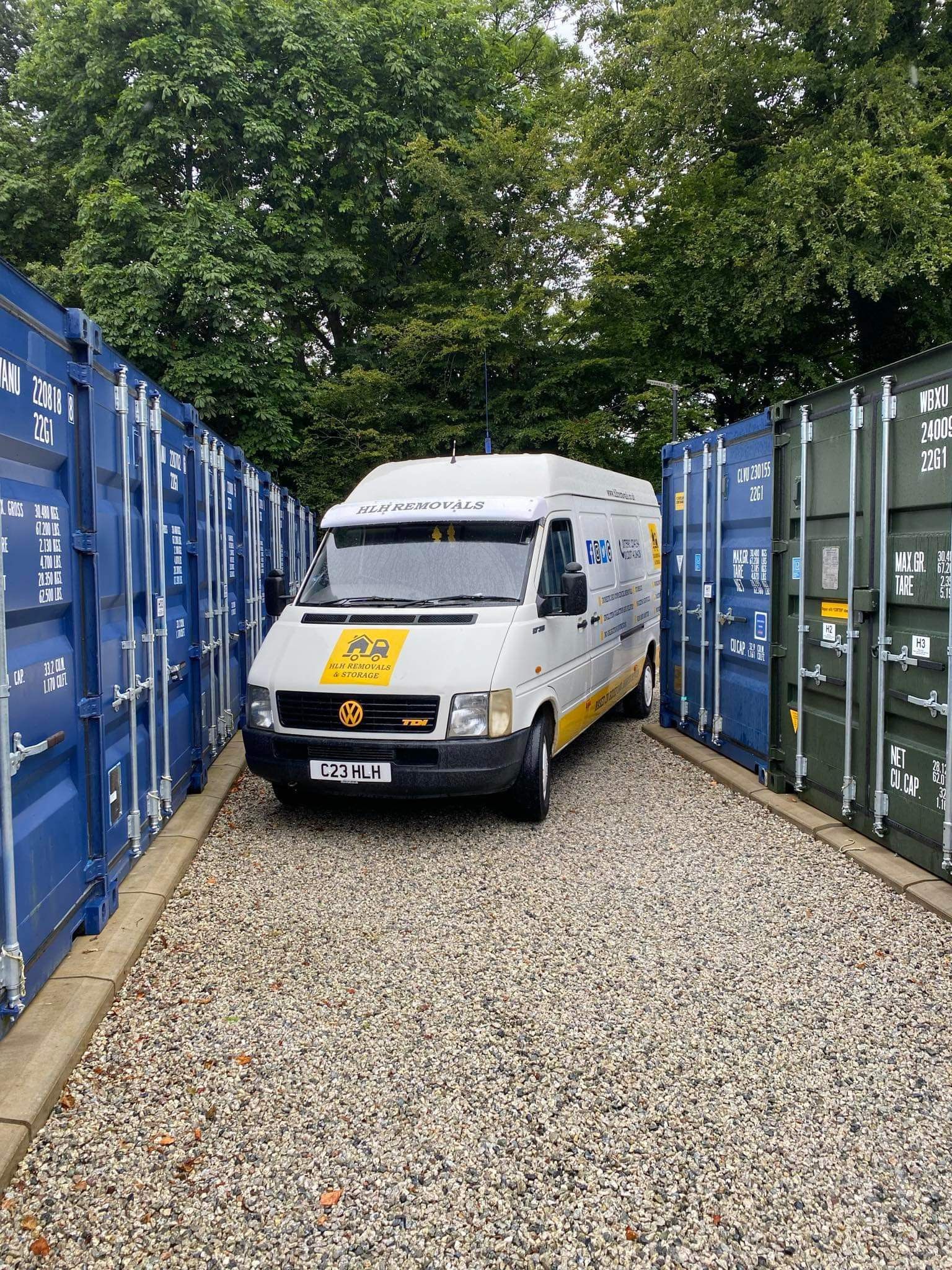 White van parked between blue and green shipping containers on gravel.
