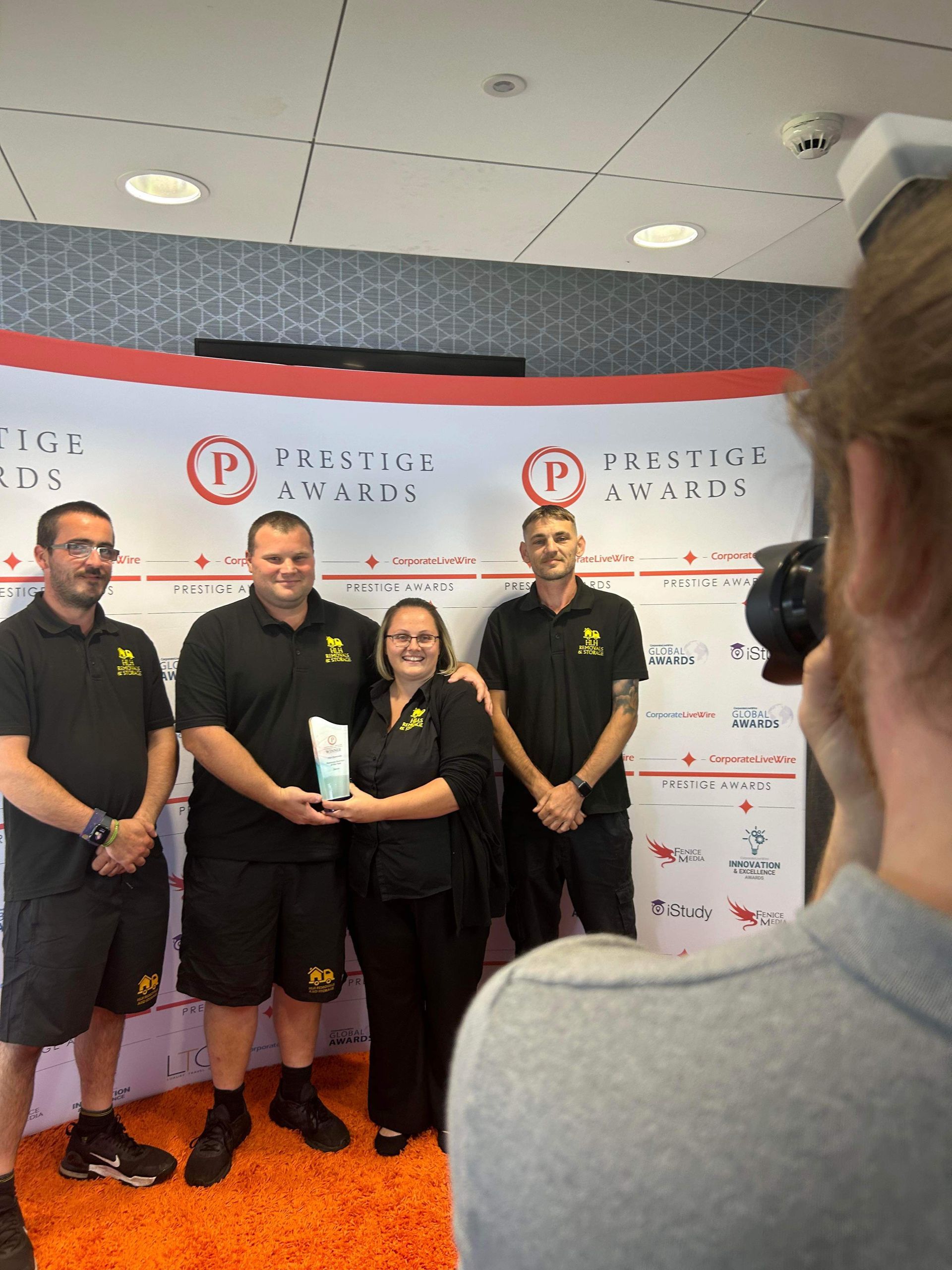 Group of four people holding an award, posed in front of a red and white banner, a photographer taking a photo.
