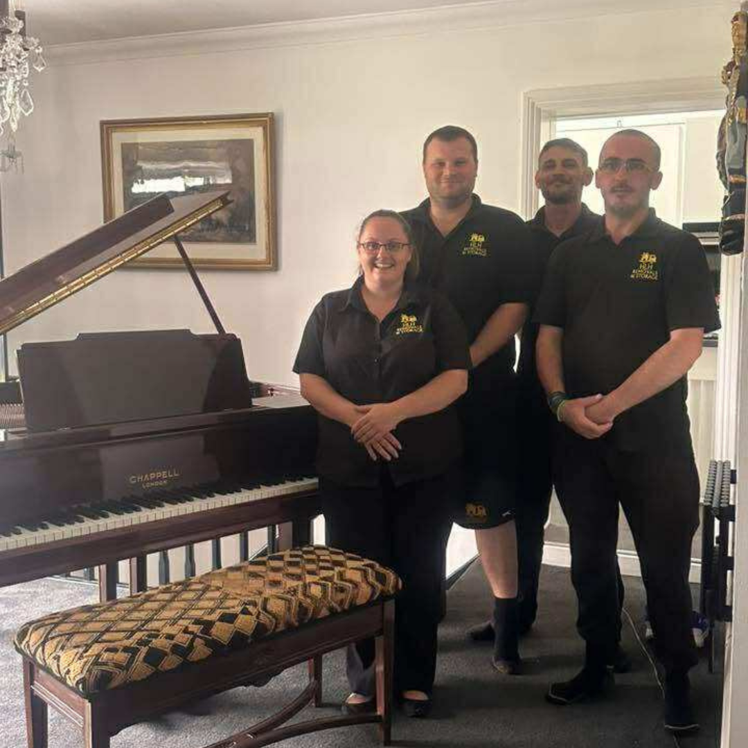 Four people in matching black shirts stand near a grand piano indoors.