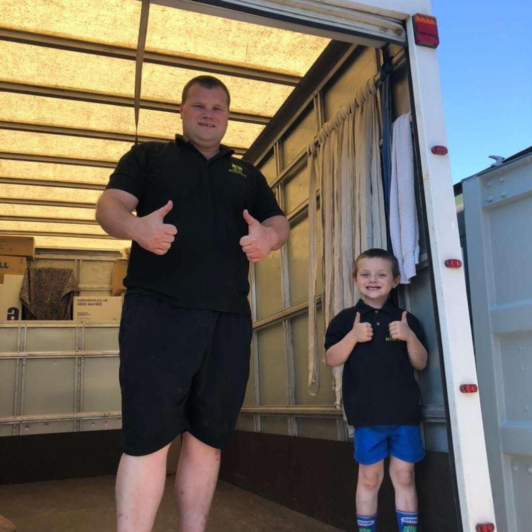 Man and boy inside a moving truck give thumbs up. Both wear black shirts, the boy blue shorts.