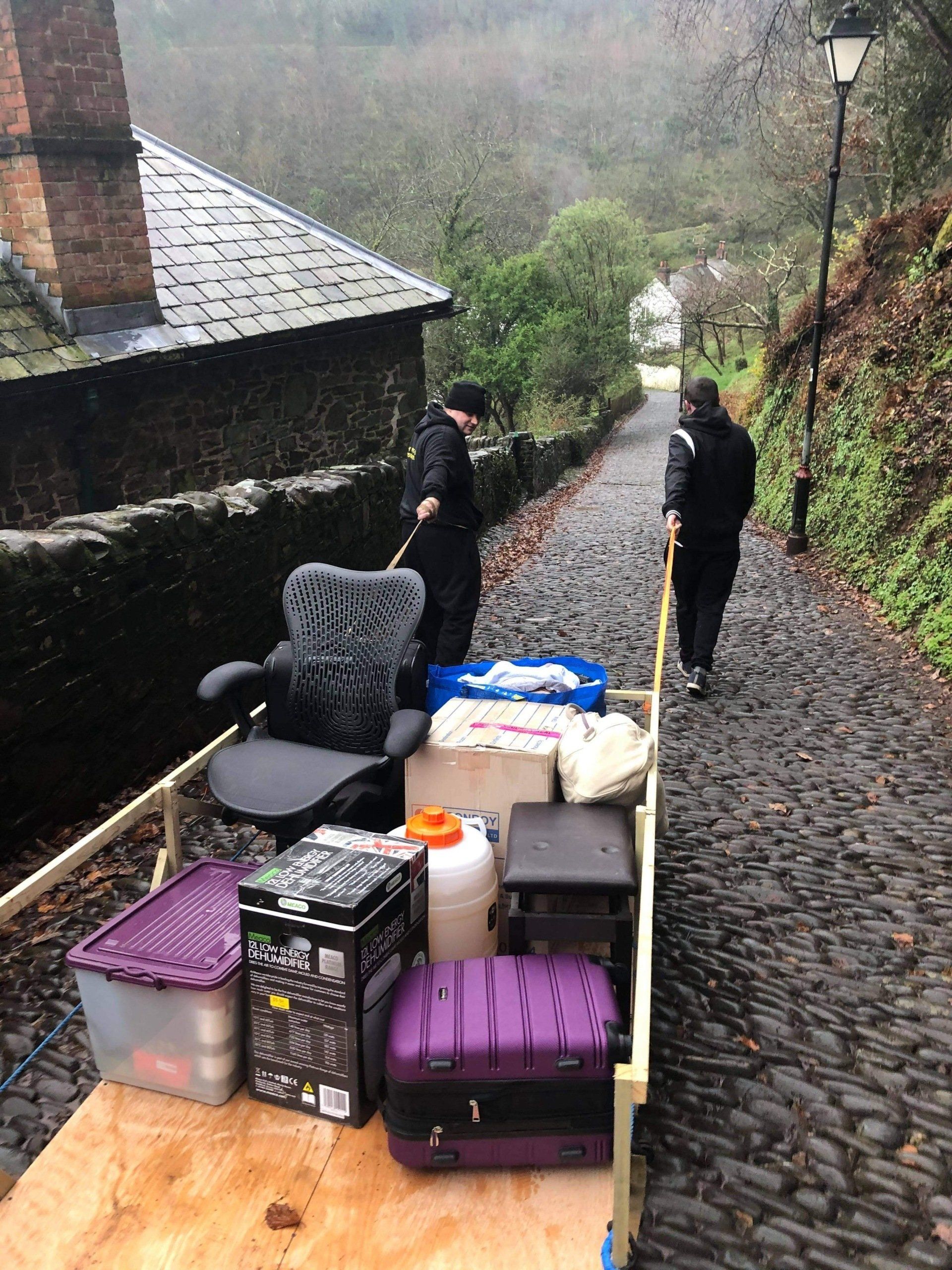 Movers using a cart to transport belongings down a narrow, cobbled street.
