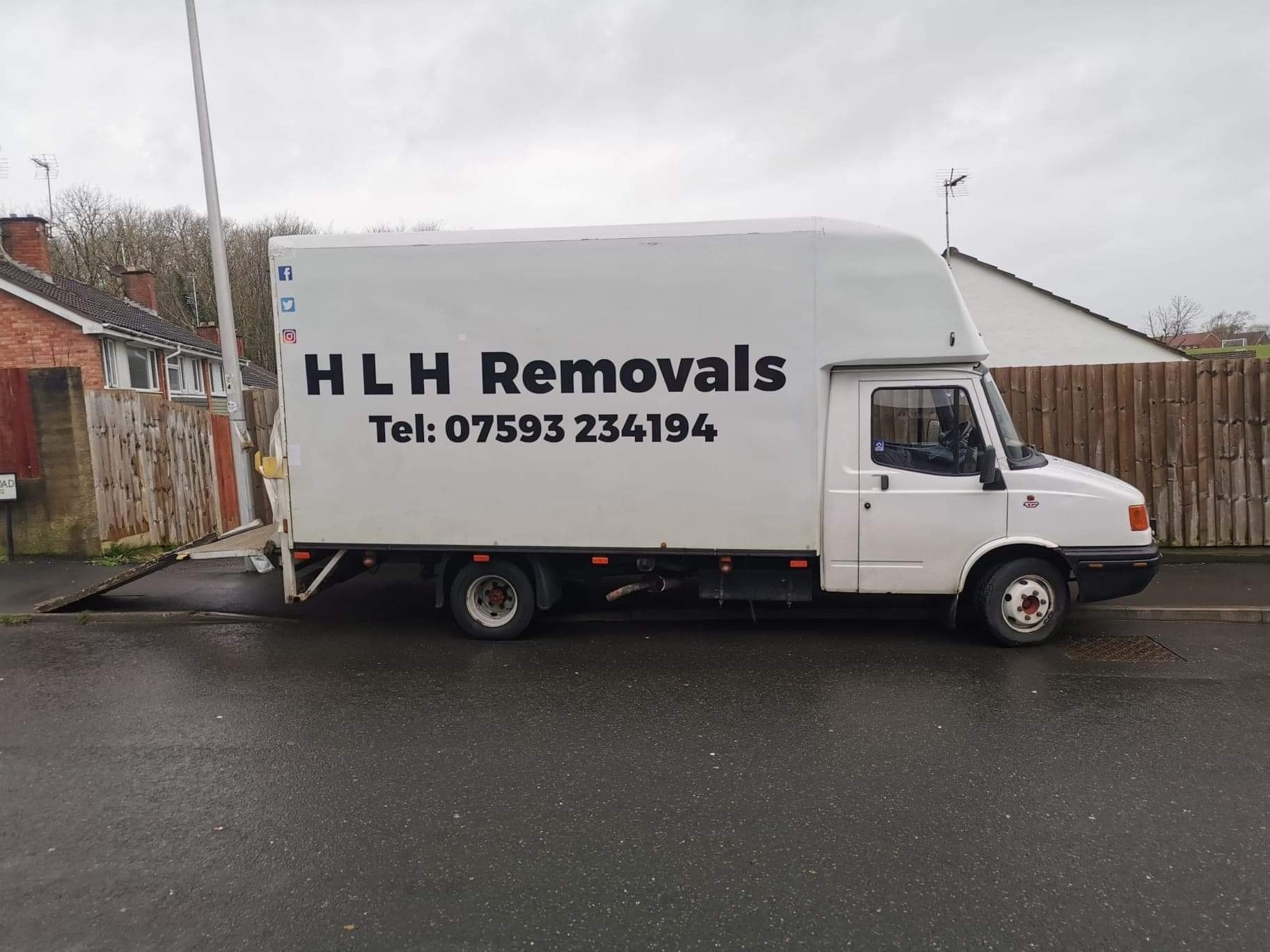 White HLH Removals truck parked on a wet street in front of houses, phone number visible.