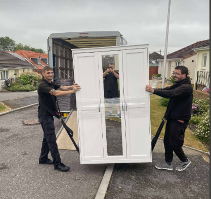 Two men carefully carry a white wardrobe off a truck in a residential area.