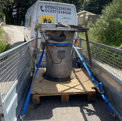 A large industrial machine secured on a wooden pallet on a trailer, blue straps, van in background.