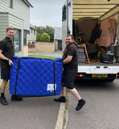 Two men in black carrying a large blue padded item from a moving truck on a street.