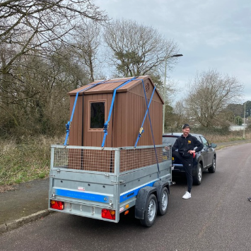 Man by trailer hauling a small brown shed on a gray trailer, parked on a road.