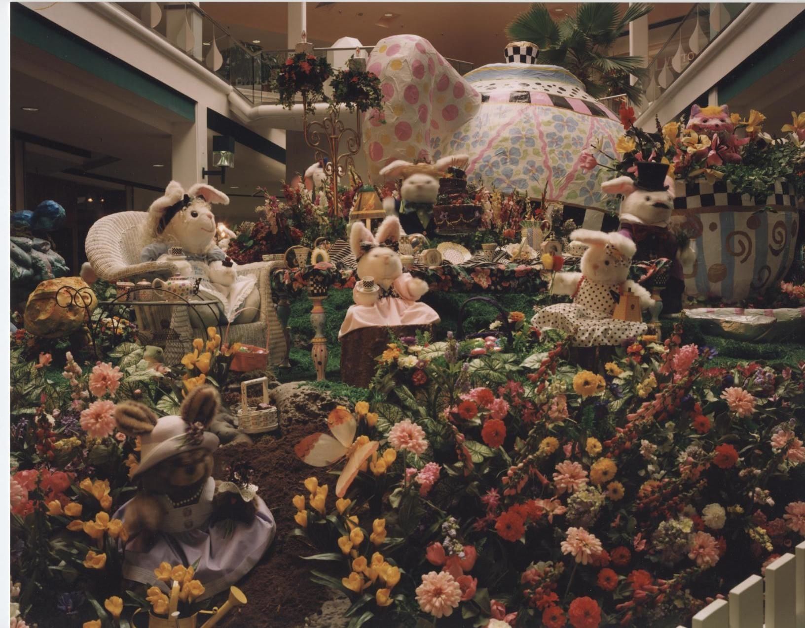 Easter display at a mall, featuring bunnies among colorful flowers, a teapot, and a tiered cake.