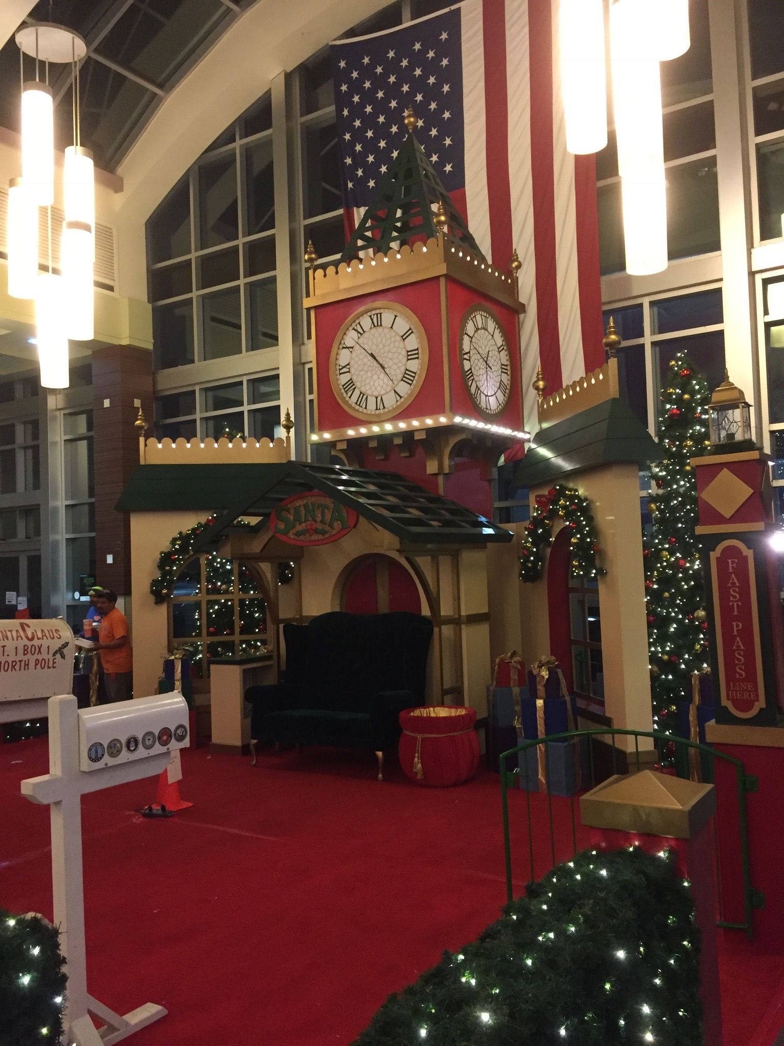 Santa's workshop display with a large clock tower, American flag backdrop, and red carpet in a mall.