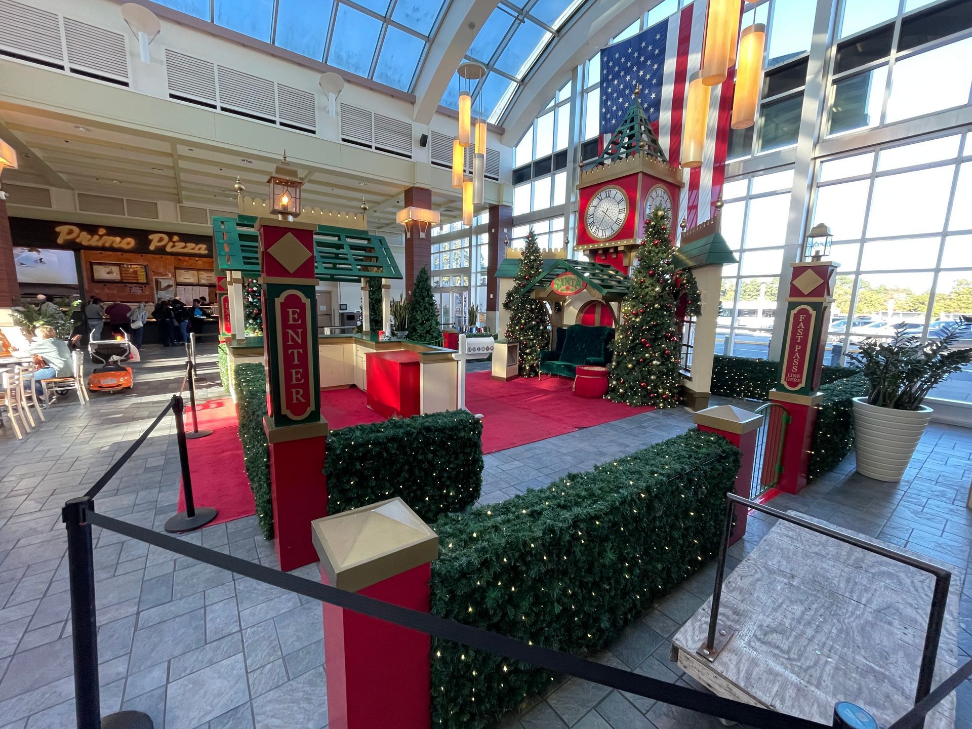Santa's workshop display in mall, red carpet, green hedge, Christmas trees, clock tower, American flag.