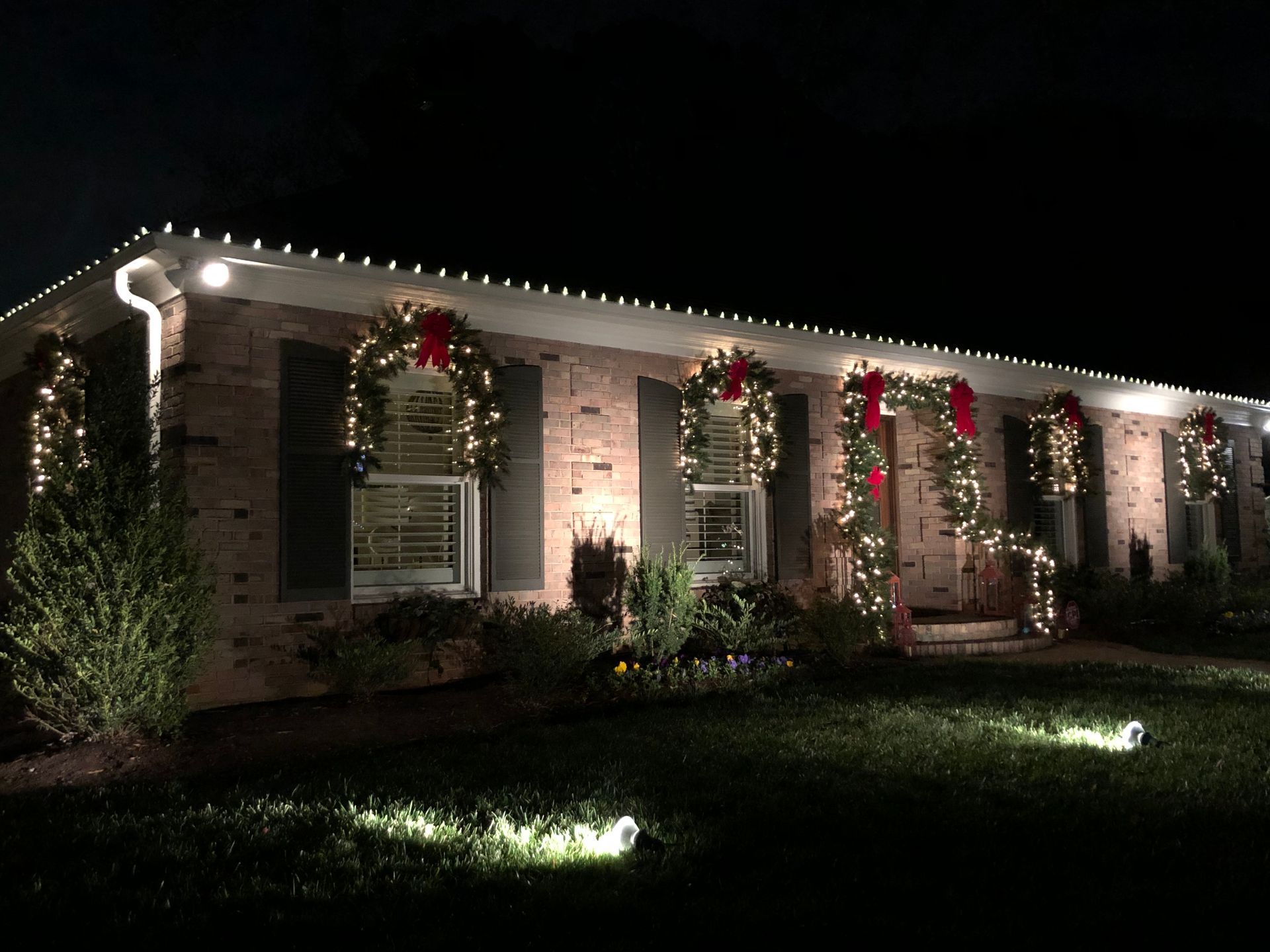 A brick house at night decorated with white Christmas lights, wreaths, and red bows.