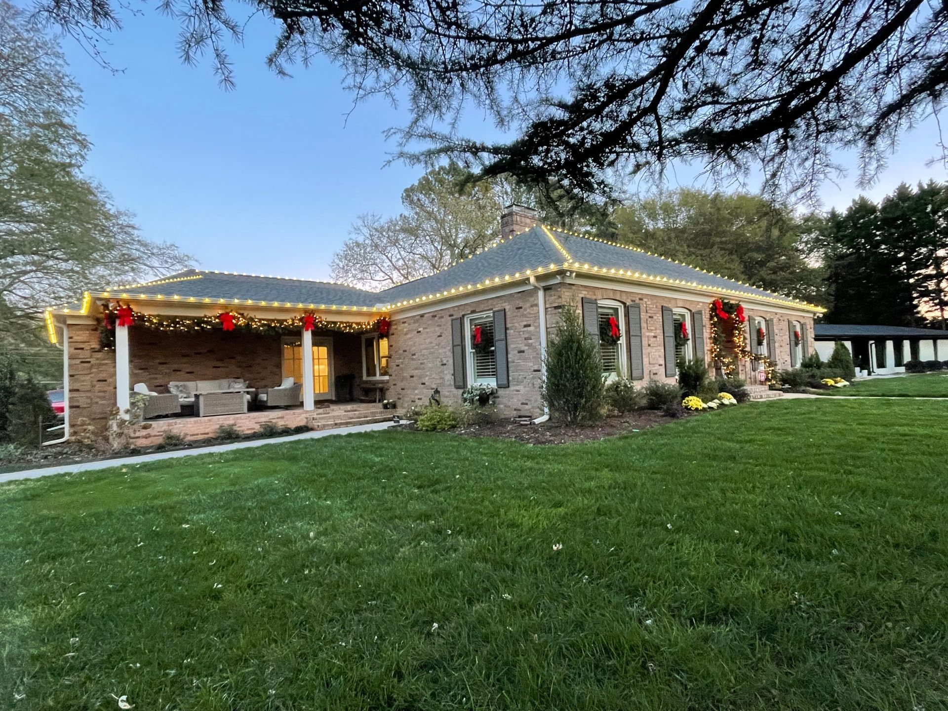 Brick house with Christmas lights and wreaths, green lawn, blue sky.