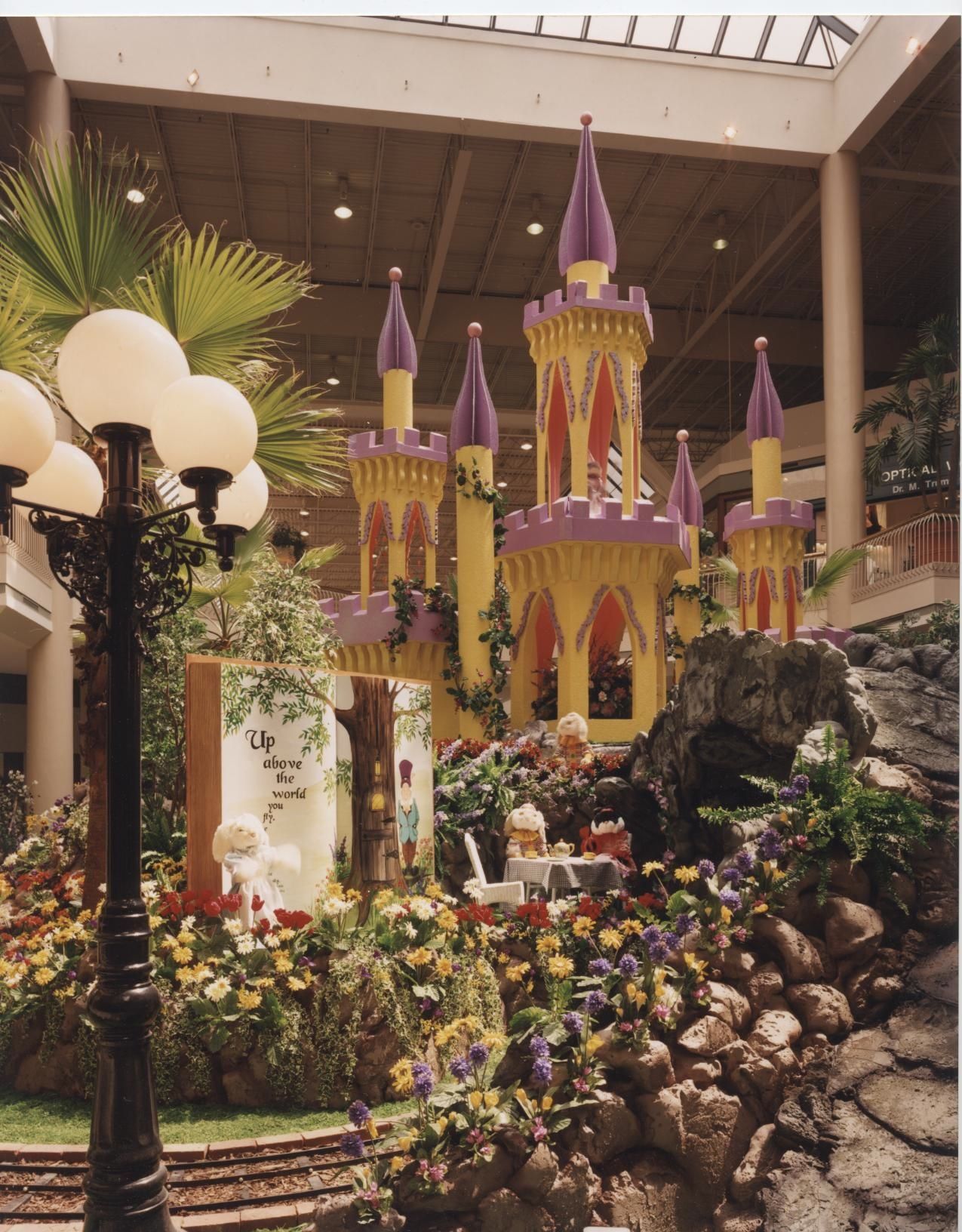 Yellow and purple fairytale castle display in a mall atrium surrounded by flowers, rocks, and a lamppost.