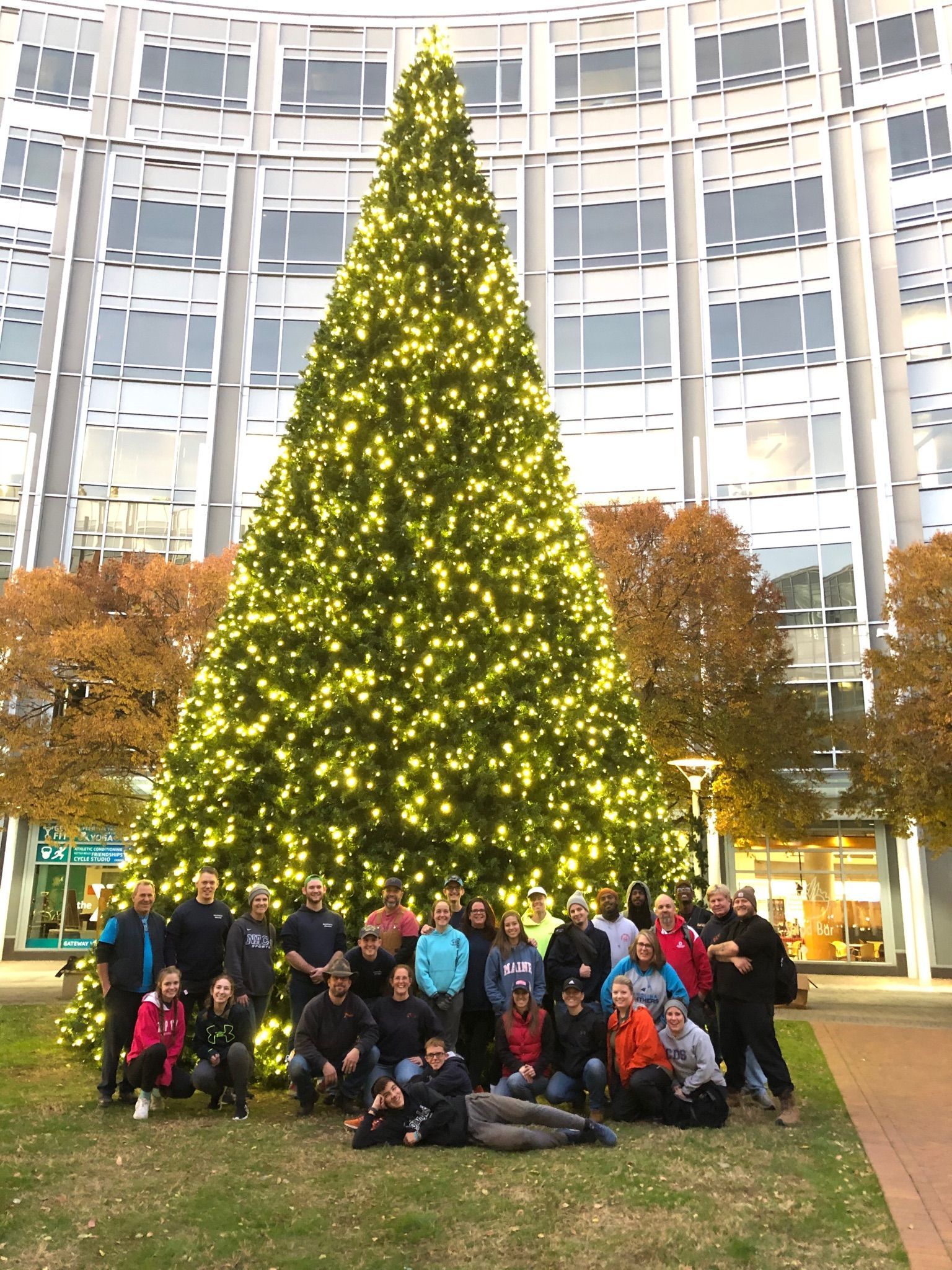 Large group posing in front of a lit Christmas tree outside a modern building.