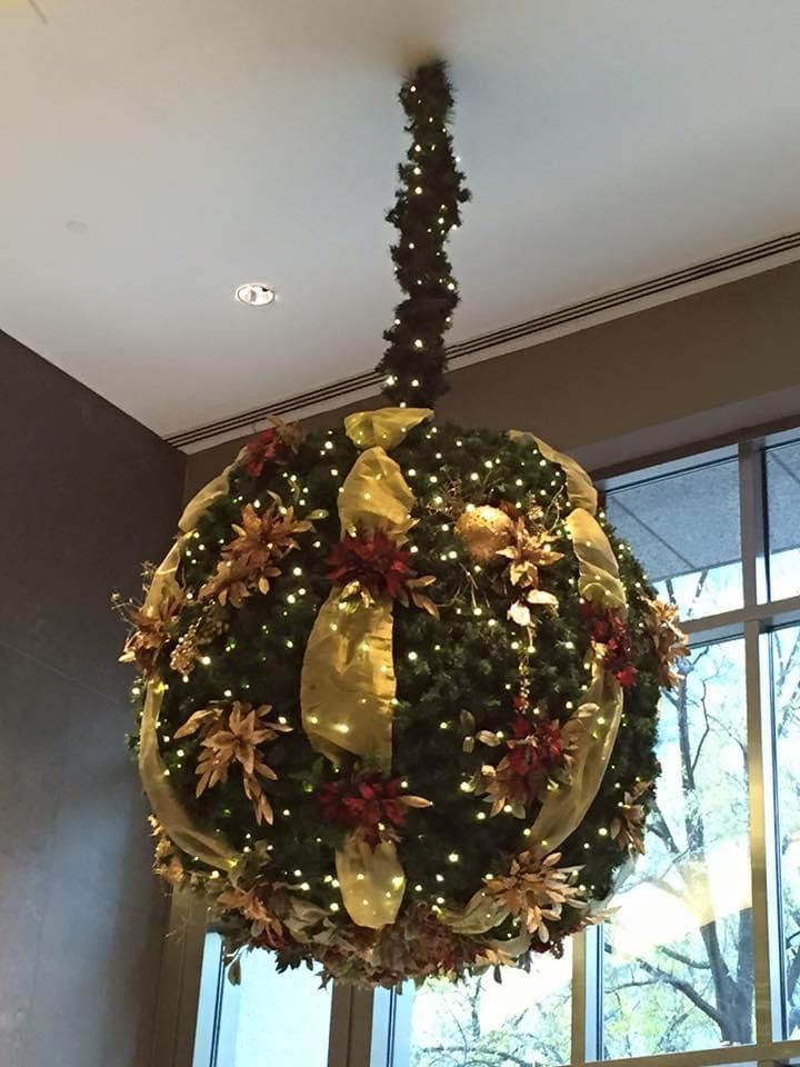 Large, spherical Christmas ornament hanging from a ceiling, decorated with lights, gold ribbons, and flowers.