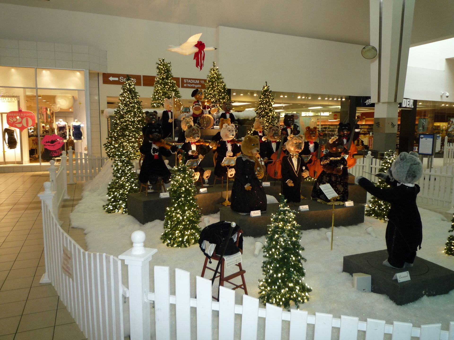 Christmas display in a mall: musicians in formal attire, trees, fake snow, conductor, and white picket fence.