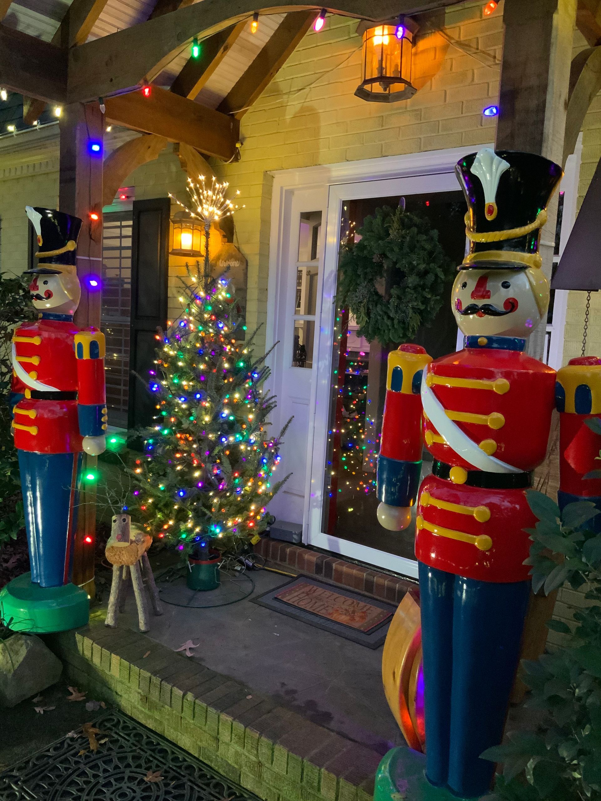 Two large toy soldiers flank a doorway with a Christmas tree and wreath, lit by colorful lights.