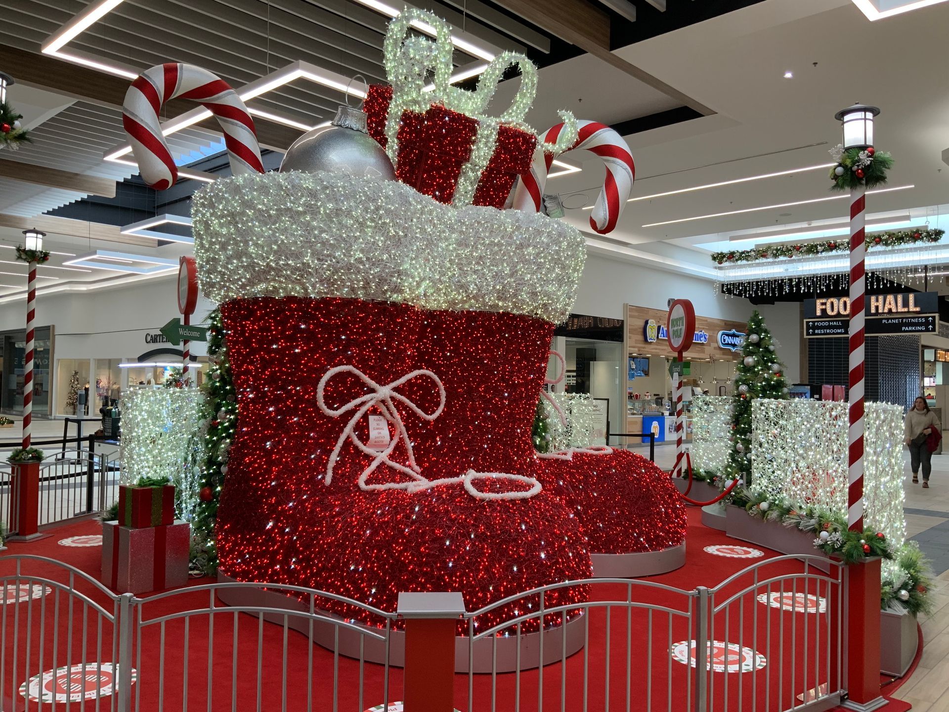 Large, sparkly red Christmas stocking display in a mall. Contains candy canes and a gift, enclosed by a red fence.