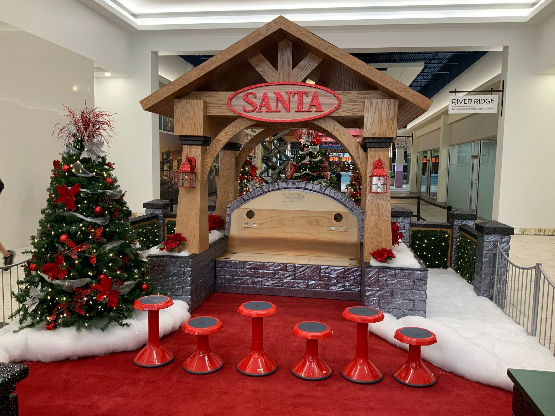 Santa's holiday display in a mall: wooden backdrop, bench, small red stools, Christmas tree, red carpet.
